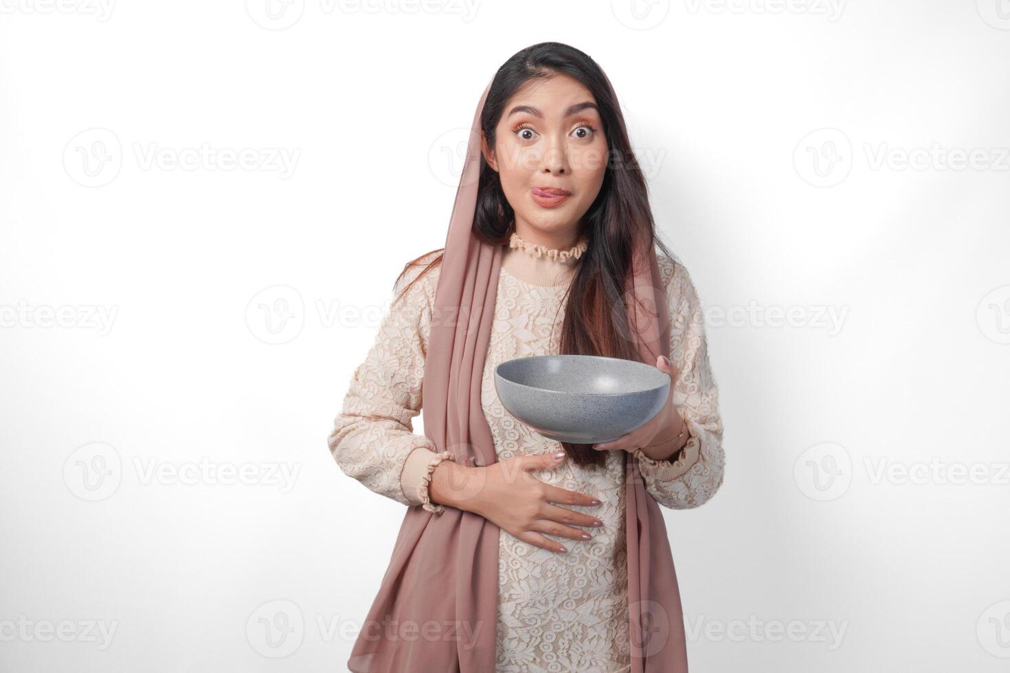 Portrait of a hungry Asian Muslim woman holding bowl thinking what to eat after fasting with copy space over the bowl, isolated white background photo