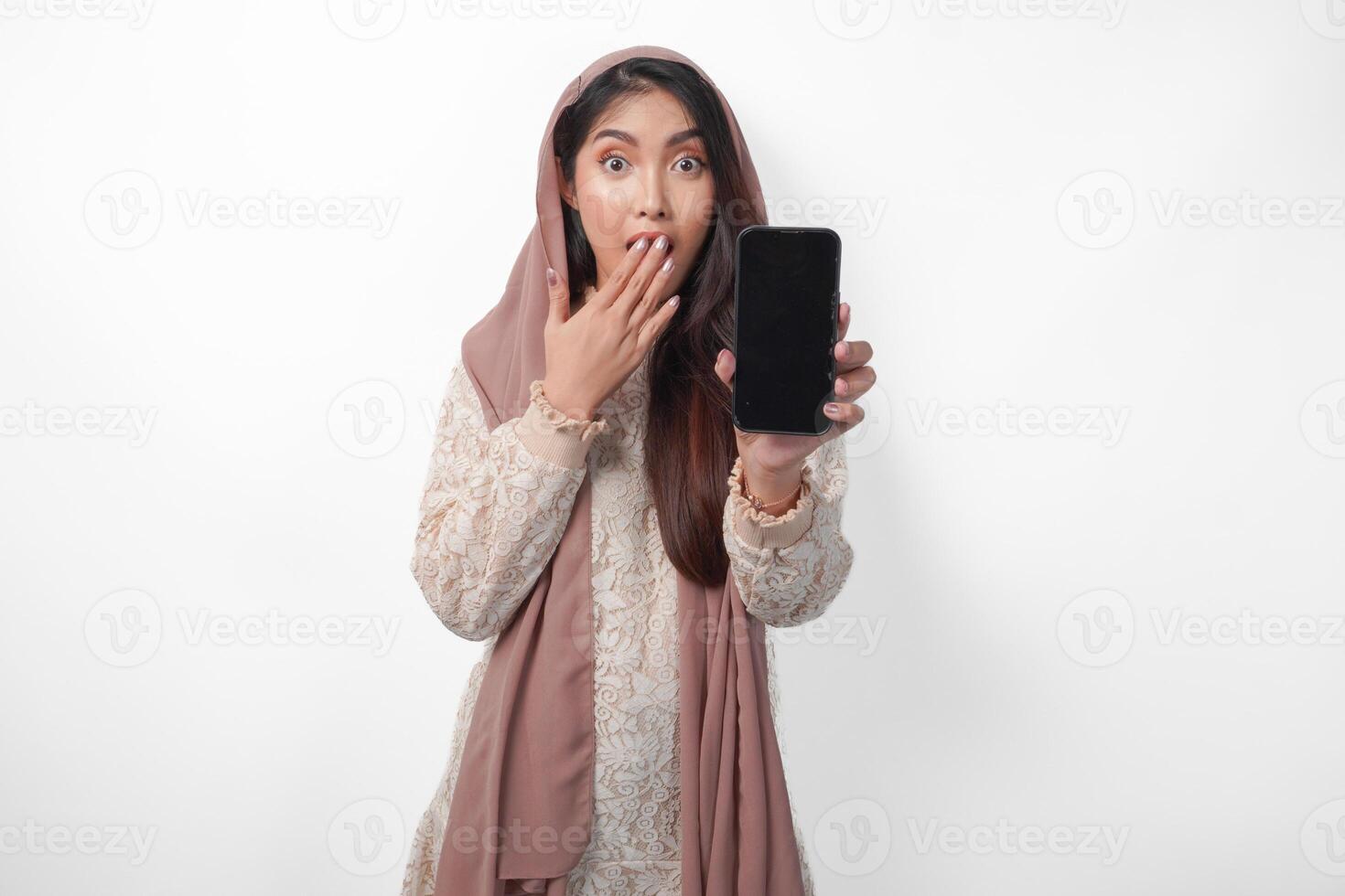 Shocked Asian Muslim woman in headscarf veil hijab with hand covering mouth while showing blank phone screen with a copy space, isolated white background photo