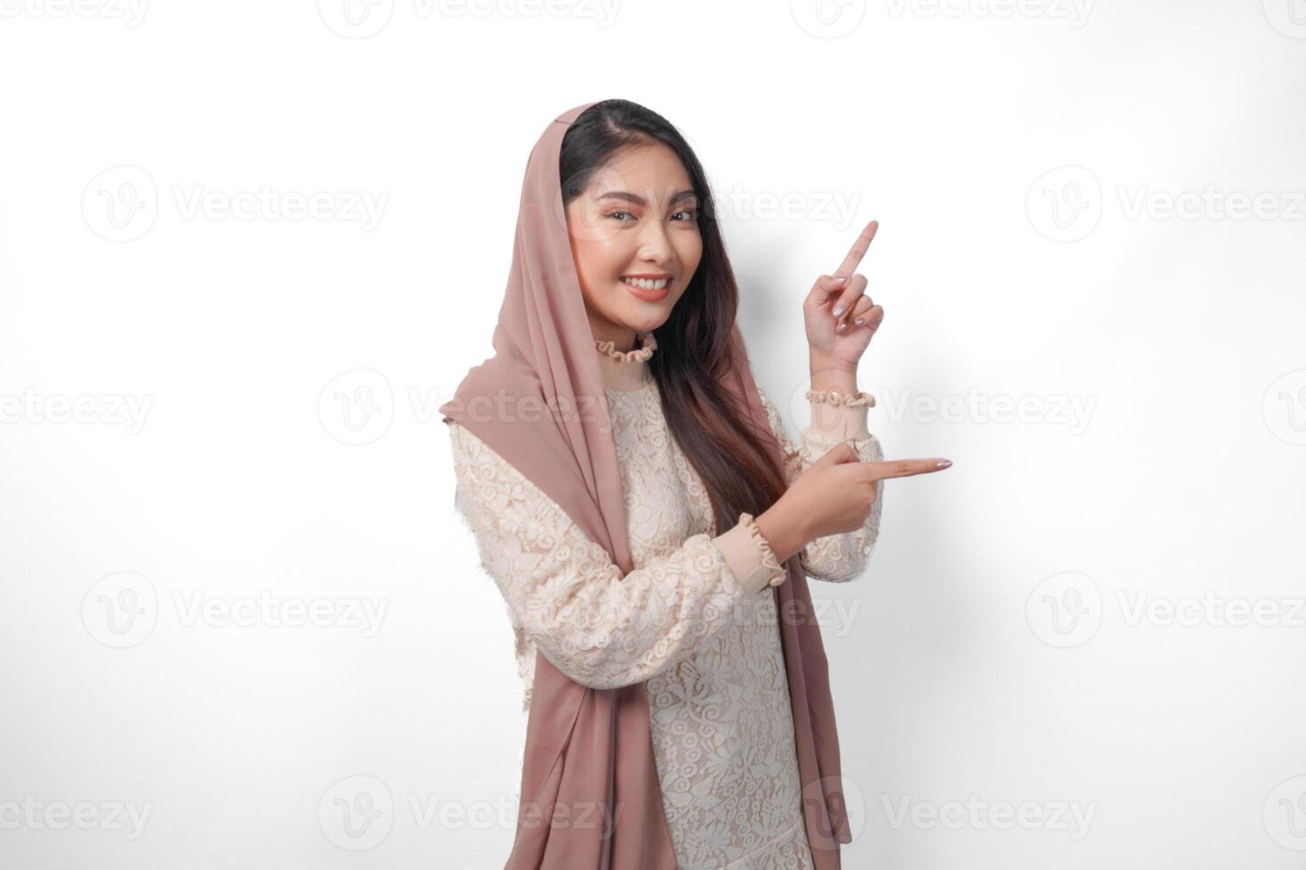 Asian Muslim woman wearing headscarf veil hijab smiling cheerfully while looking and pointing to copy space beside her, standing on isolated white background photo
