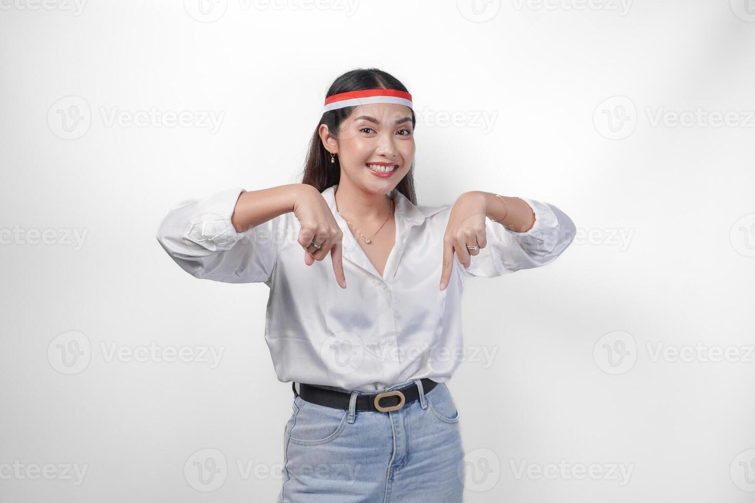 Happy Asian woman wearing Indonesia flag colored headband and casual outfit while smiling cheerfully and pointing down at the copy space. Independence day advertisement concept photo