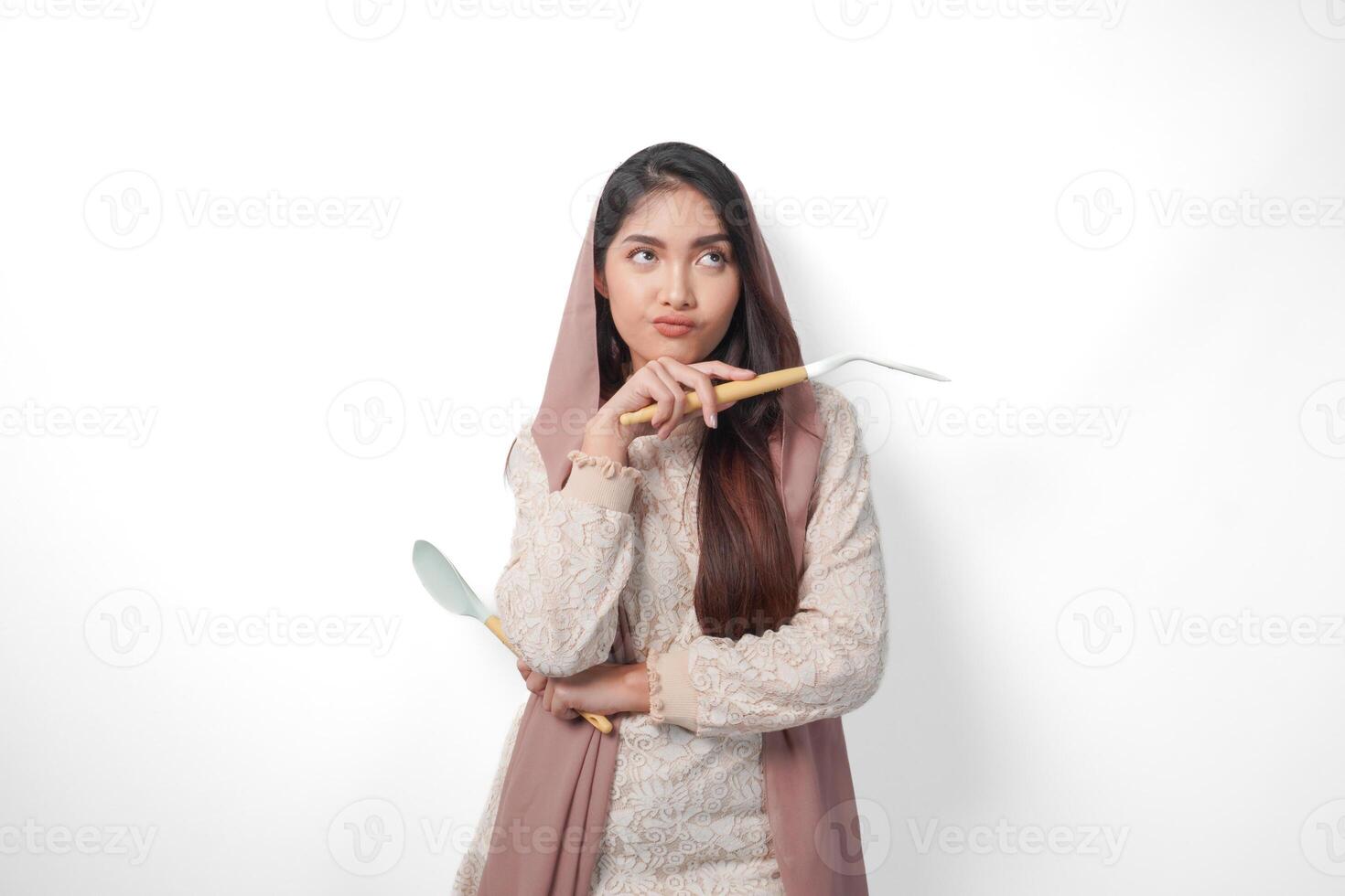 pensativo asiático musulmán mujer vistiendo Pañuelo velo hijab enfocado y mirando a Copiar espacio junto a su mientras participación espátula o cocina utensilios, en pie en aislado blanco antecedentes foto