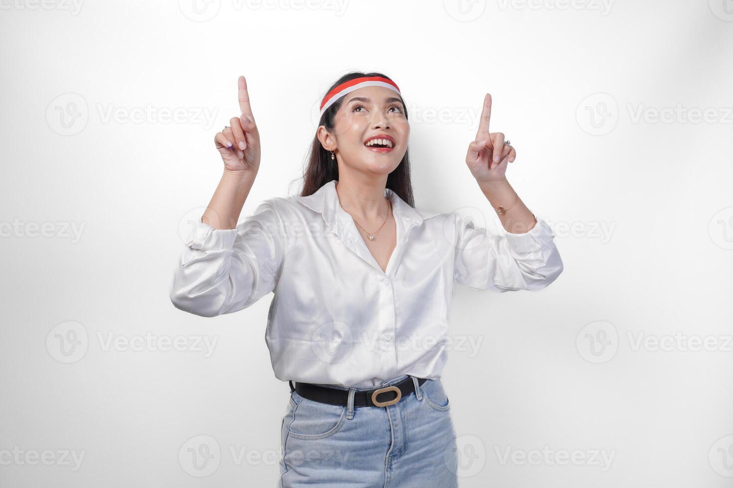 Happy Asian woman wearing Indonesia flag colored headband and casual outfit while smiling cheerfully and pointing up the copy space upwards. Independence day advertisement concept photo