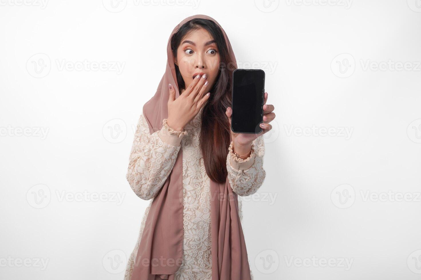 Shocked Asian Muslim woman in headscarf veil hijab with hand covering mouth while showing blank phone screen with a copy space, isolated white background photo