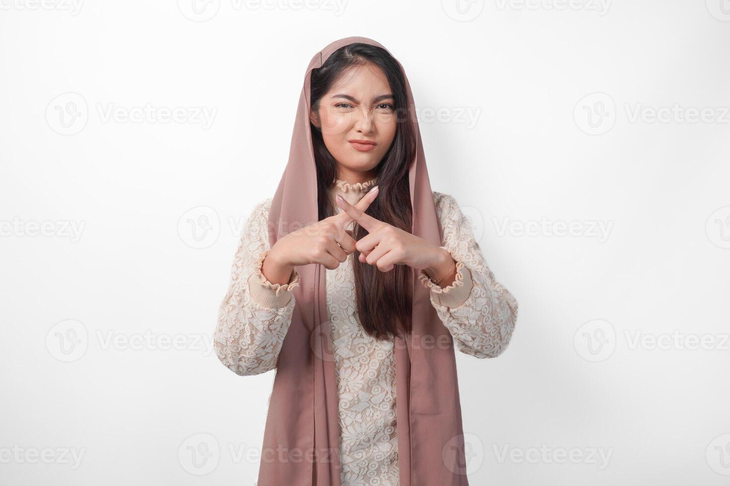 Young Asian Muslim woman wearing veil hijab gesturing a no rejection, refuse using hands and fingers, isolated by white background. Ramadan and Eid Mubarak concept photo