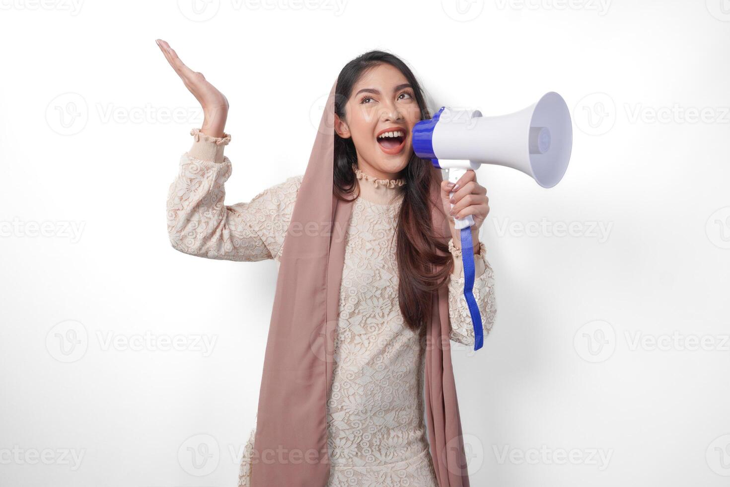 Young Asian muslim woman wearing headscarf veil hijab shouting at megaphone while pointing up to the copy space upwards, isolated on white background studio. Ramadan and Eid Mubarak concept. photo