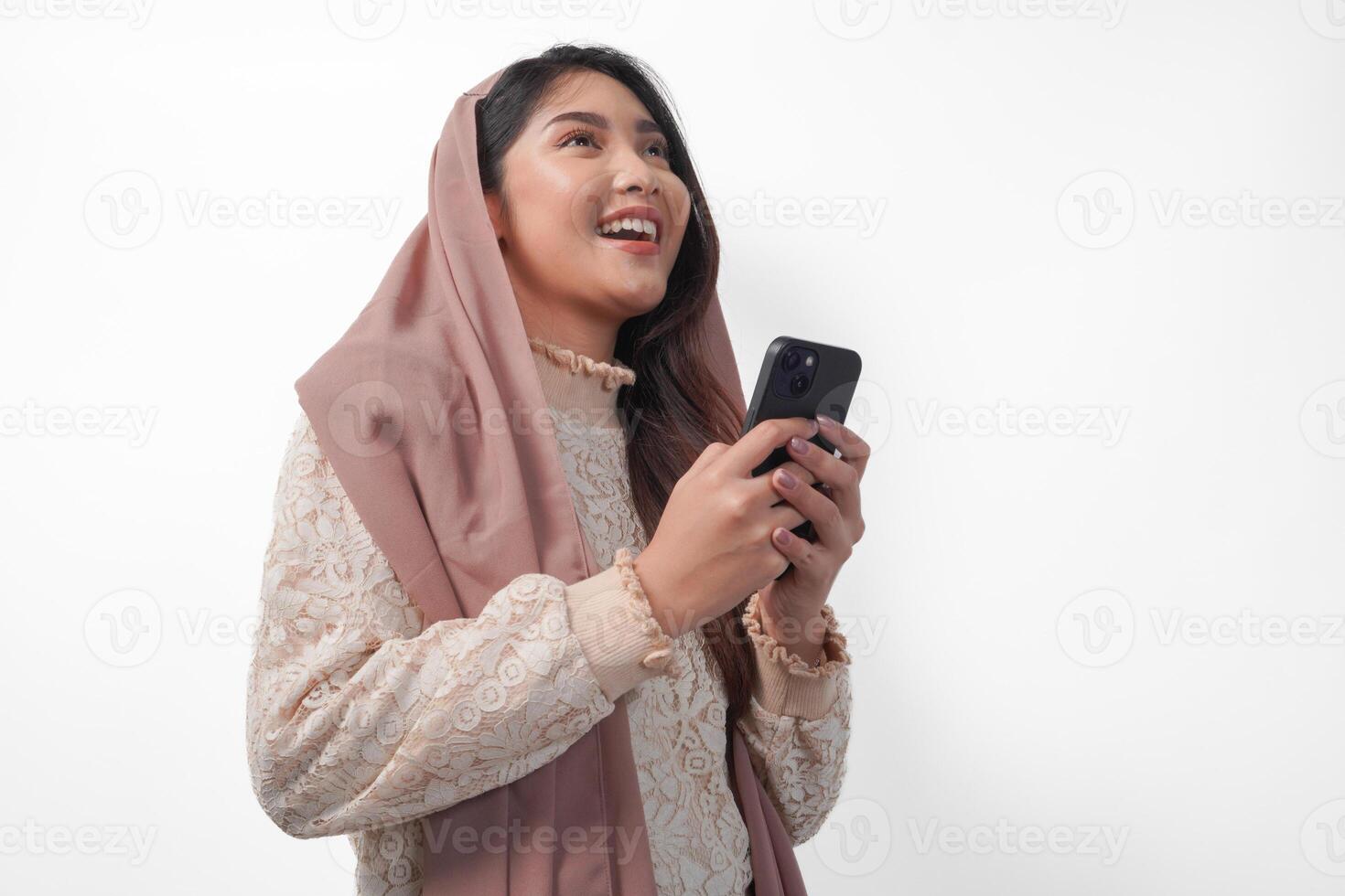 Cheerful young Asian Muslim woman in veil hijab smiling and feeling happy while holding a smartphone from side view. Ramadan and Eid Mubarak concept photo