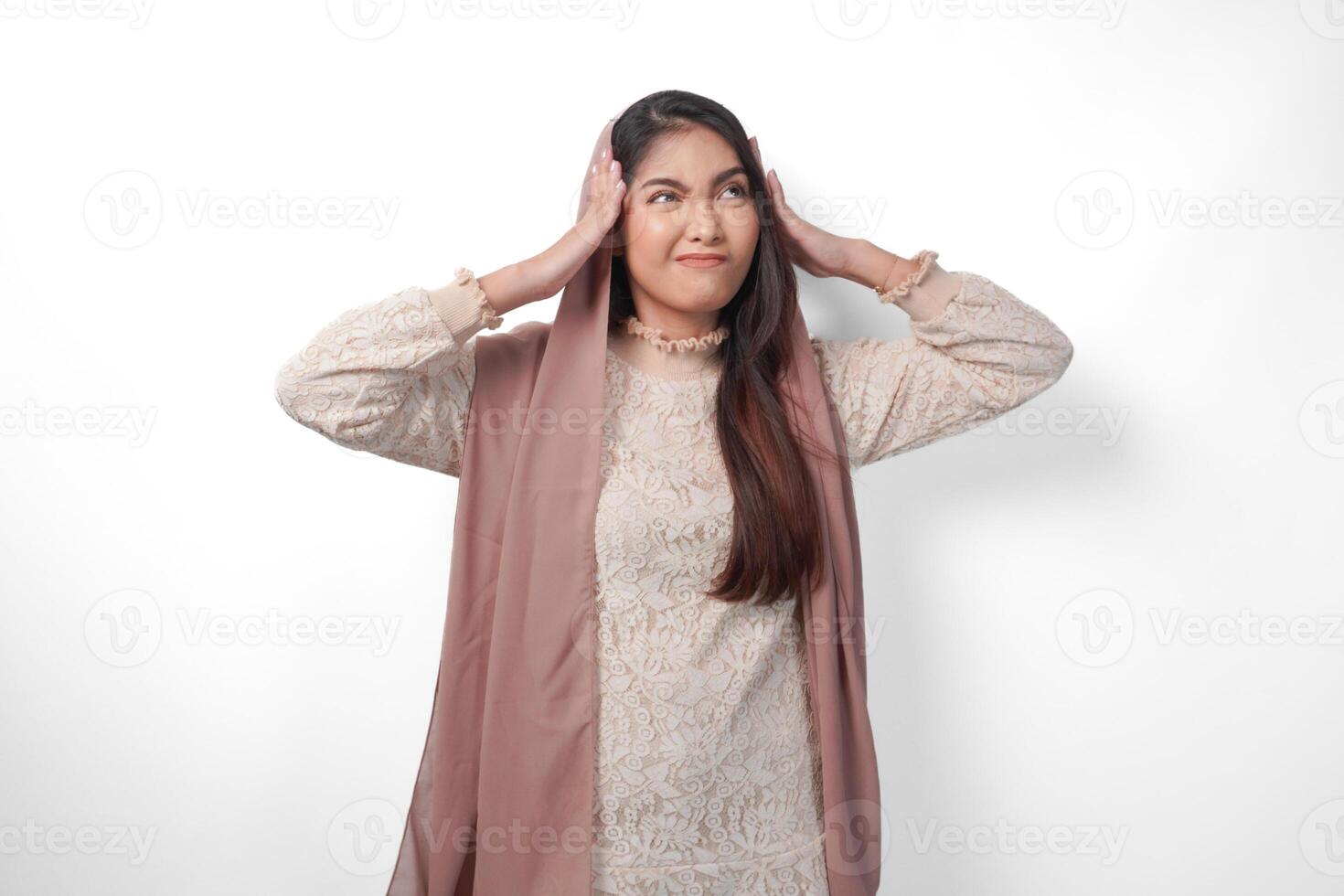 An Asian Muslim woman raising both hands to hold her head feeling stressed and overwhelmed, crying and screaming desperately, isolated white background photo