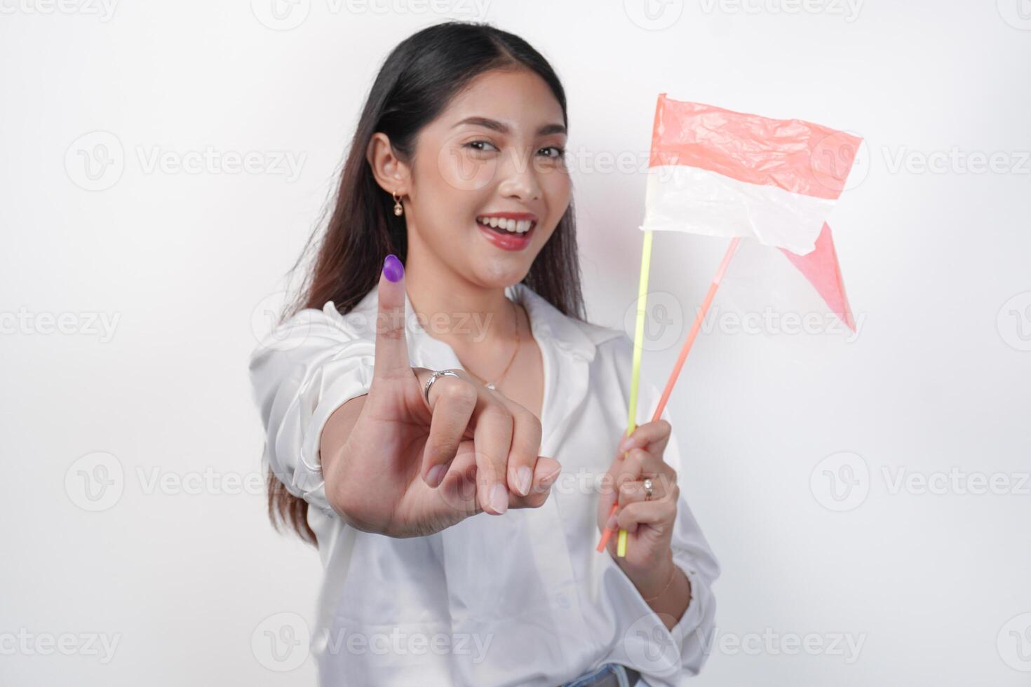 Portrait of happy Asian woman proudly showing little finger dipped in purple ink after voting for president and parliament election, left hand holding Indonesia mini flags. Independence day concept. photo