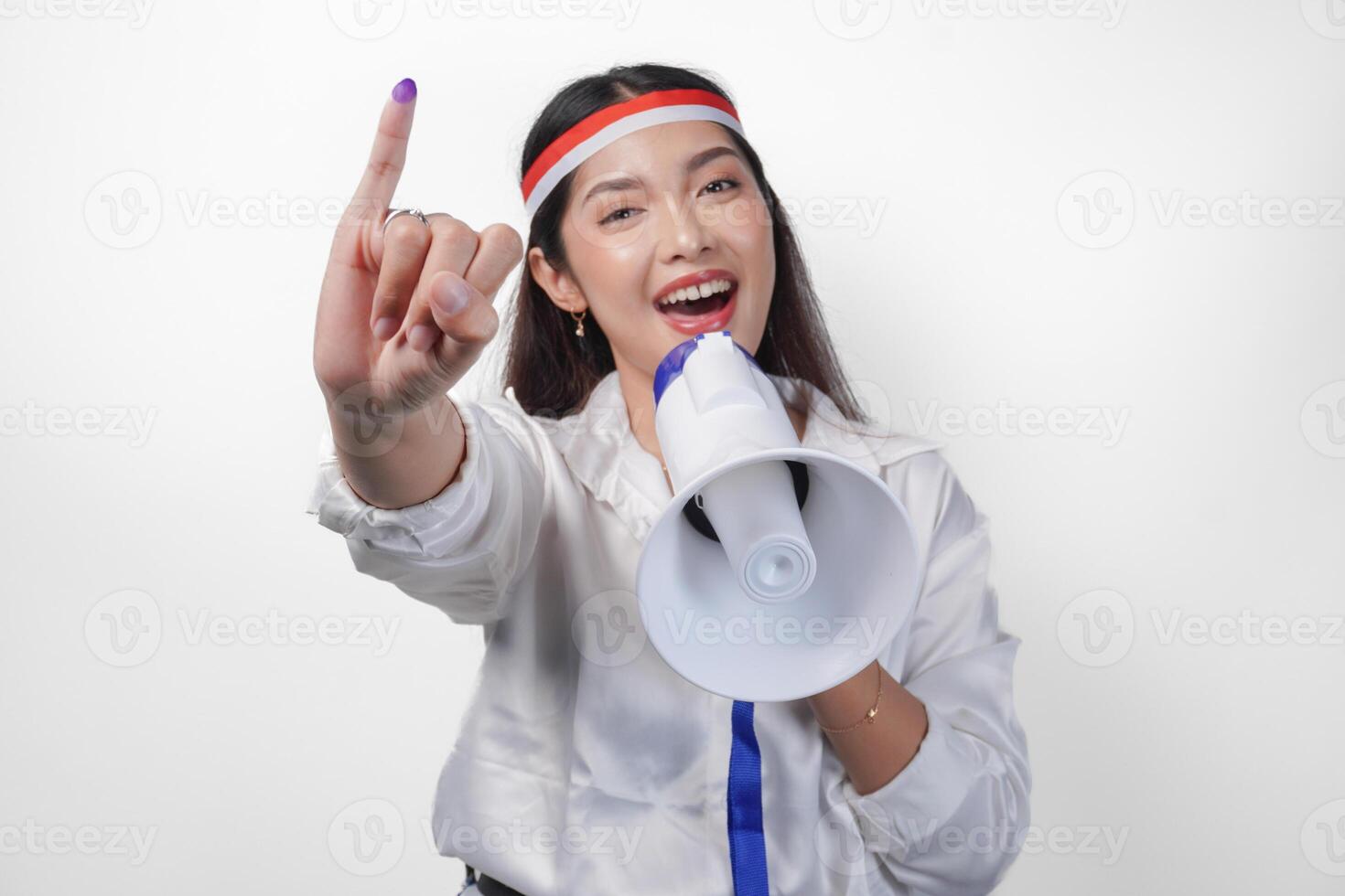 Energetic Indonesian woman in casual formal outfit wearing country flag headband while holding and shouting at megaphone, proudly showing little finger dipped in purple ink after voting for election photo