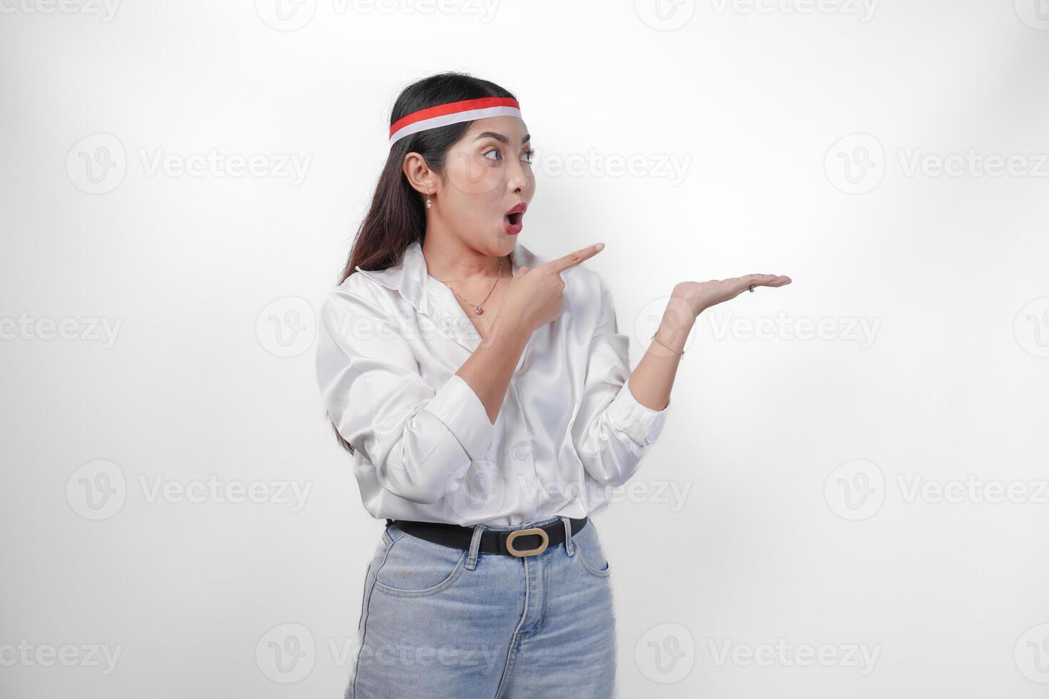 Shocked Asian woman wearing white shirt and flag headband presenting and pointing at the copy space on the side, standing on isolated white background. Independence day advertisement concept photo