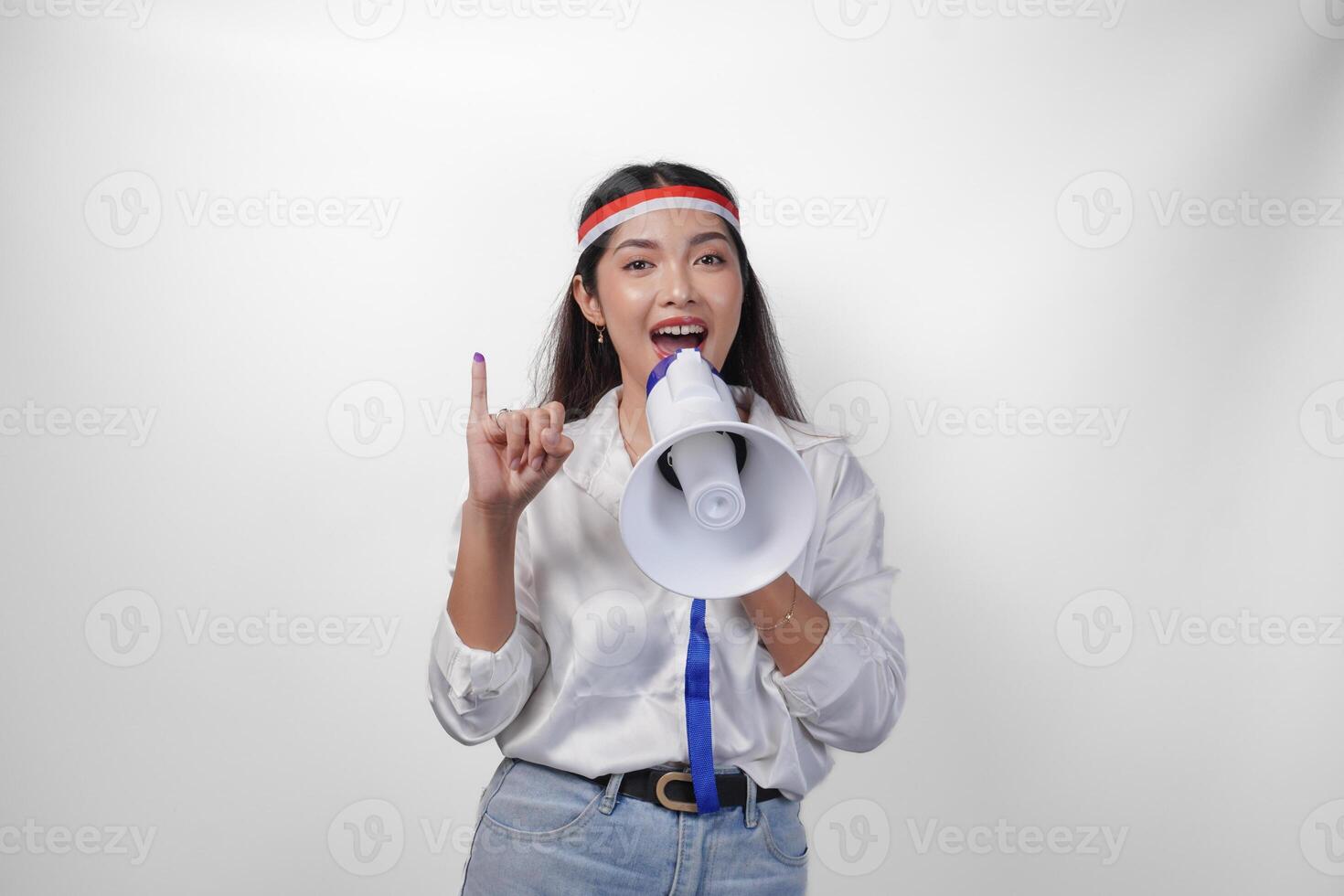 Energetic Indonesian woman in casual formal outfit wearing country flag headband while holding and shouting at megaphone, proudly showing little finger dipped in purple ink after voting for election photo