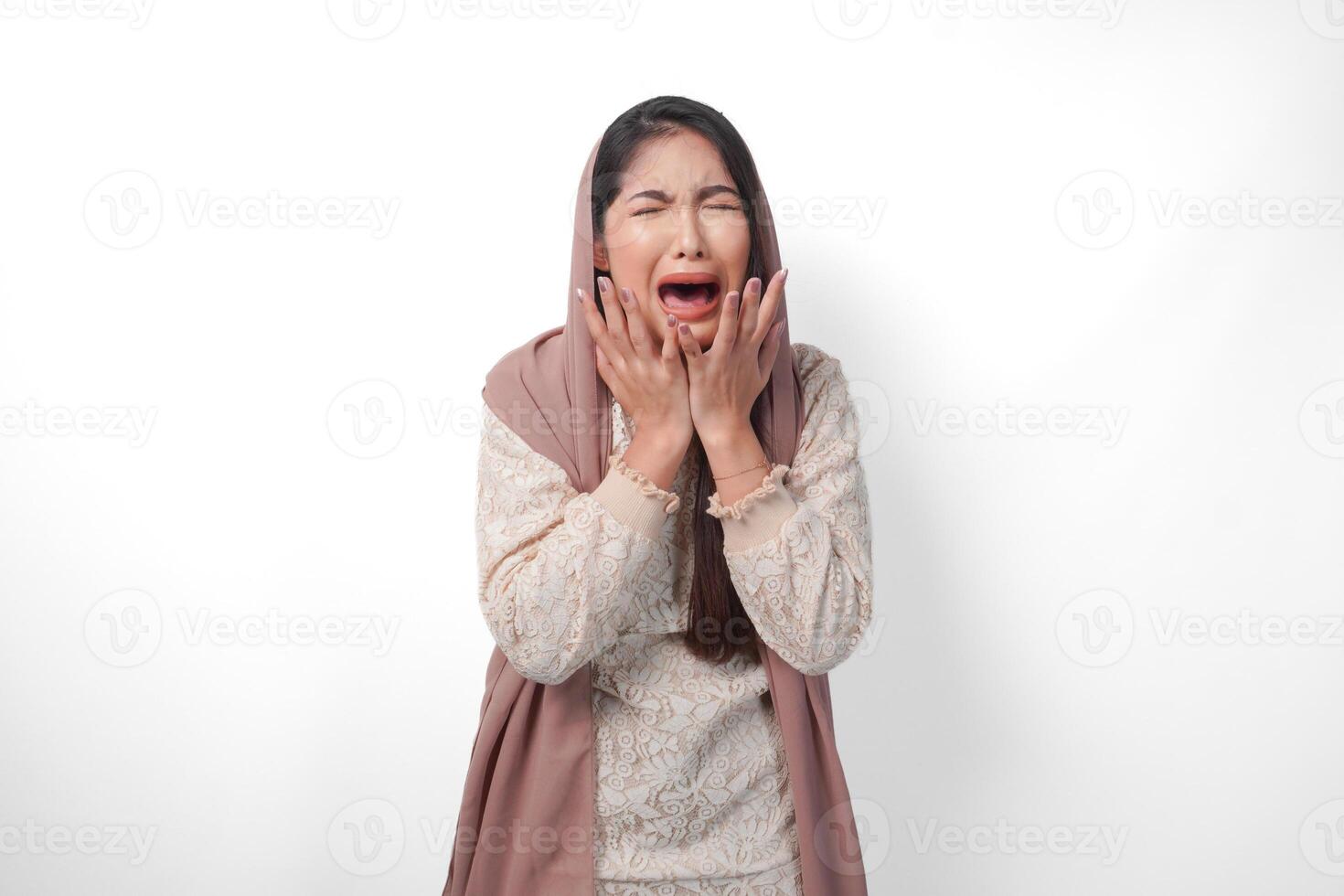 An Asian Muslim woman raising both hands to hold her head feeling stressed and overwhelmed, crying and screaming desperately, isolated white background photo