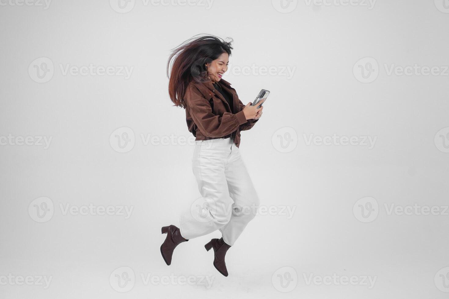 alegre asiático mujer saltando y sonriente en aire mientras participación y mirando a teléfono, sensación emocionado con un amplio risa en aislado blanco antecedentes foto