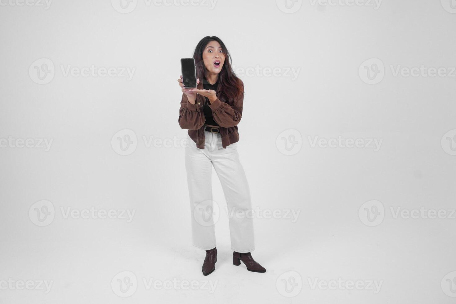 Portrait of a shocked Asian woman wearing casual outfit holding and showing her phone while her mouth wide open, standing on isolated white background photo
