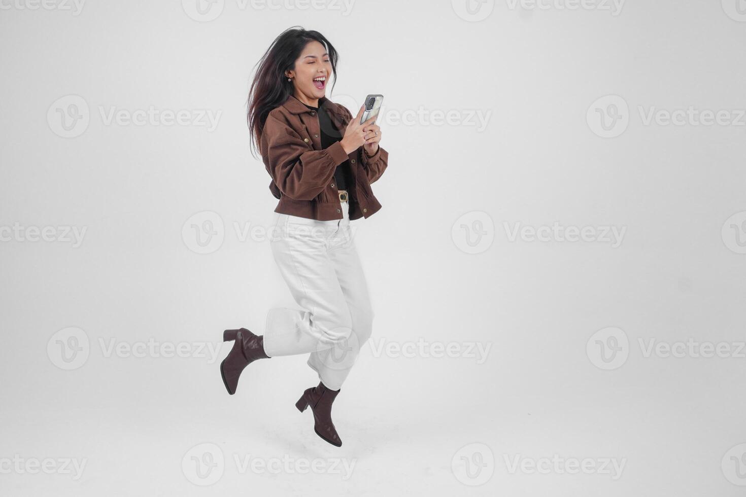 alegre asiático mujer saltando y sonriente en aire mientras participación y mirando a teléfono, sensación emocionado con un amplio risa en aislado blanco antecedentes foto