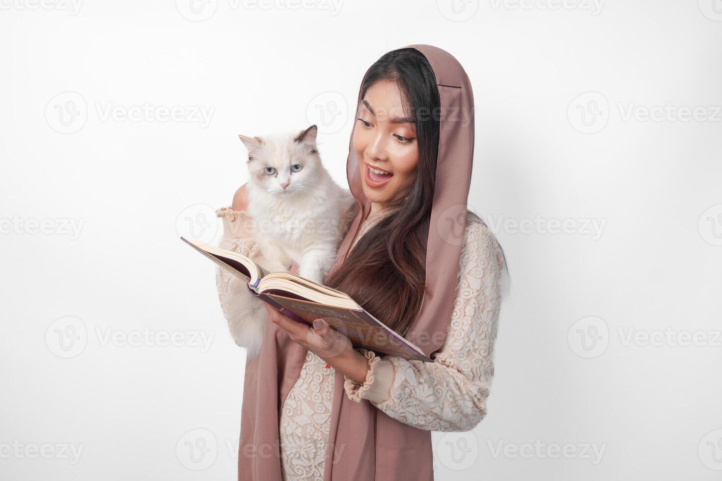 Attractive young Asian Muslim woman in veil hijab smiling while hugging a white ragdoll cat pet and holding Al Quran on the other hand photo