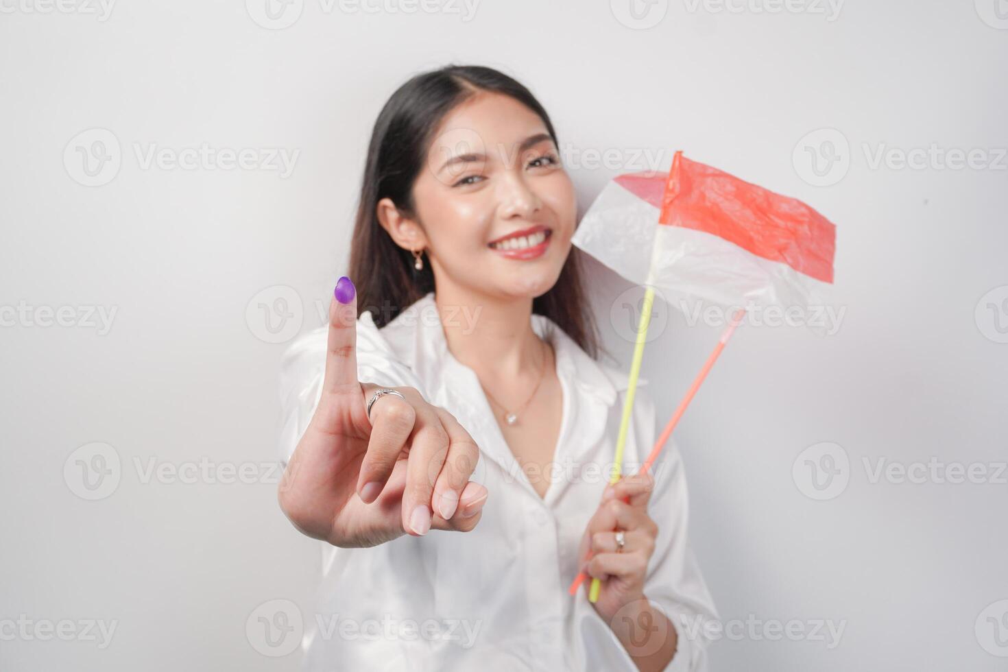 Portrait of happy Asian woman proudly showing little finger dipped in purple ink after voting for president and parliament election, left hand holding Indonesia mini flags. Independence day concept. photo