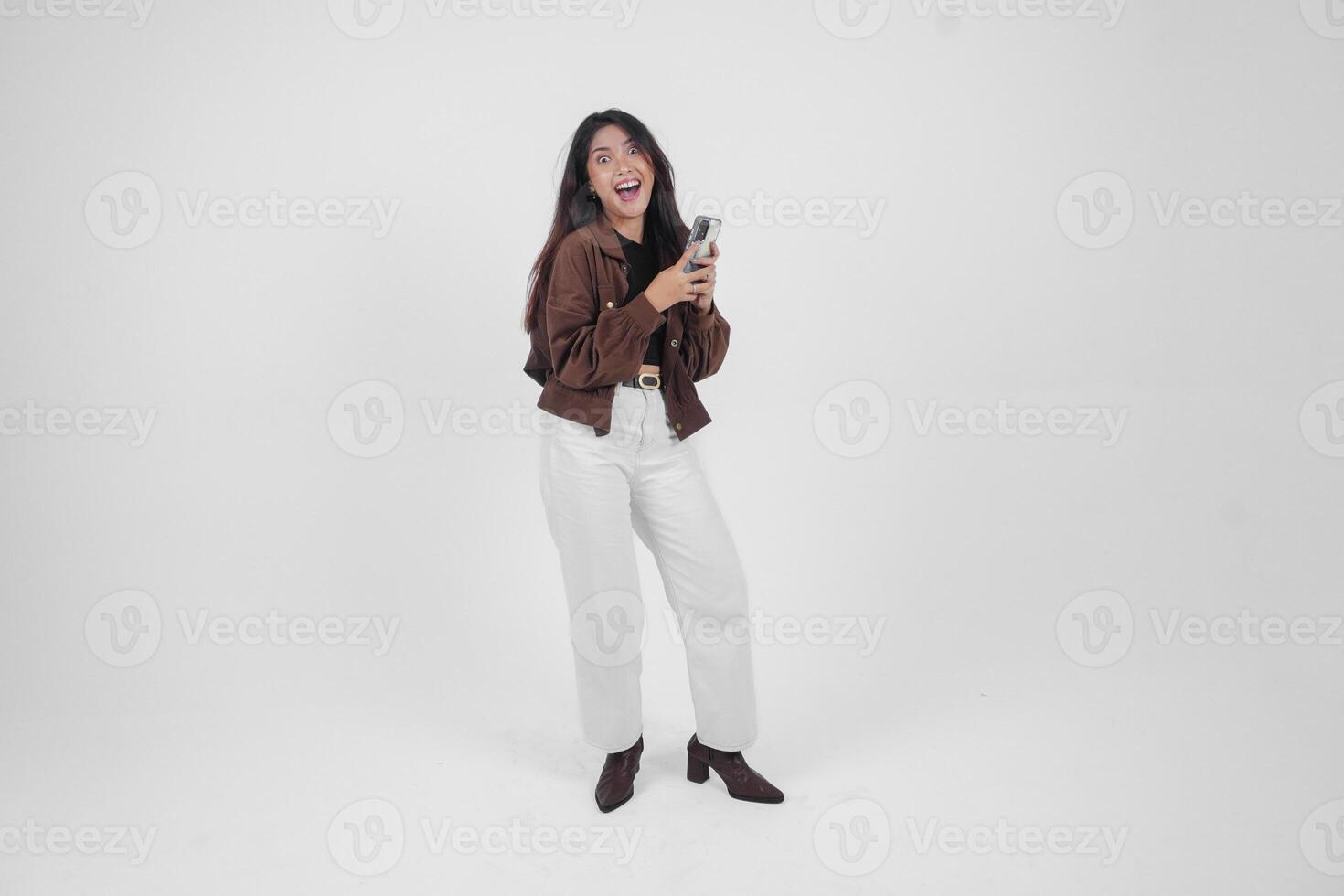 Portrait of a shocked Asian woman wearing casual outfit holding and showing her phone while her mouth wide open, standing on isolated white background photo