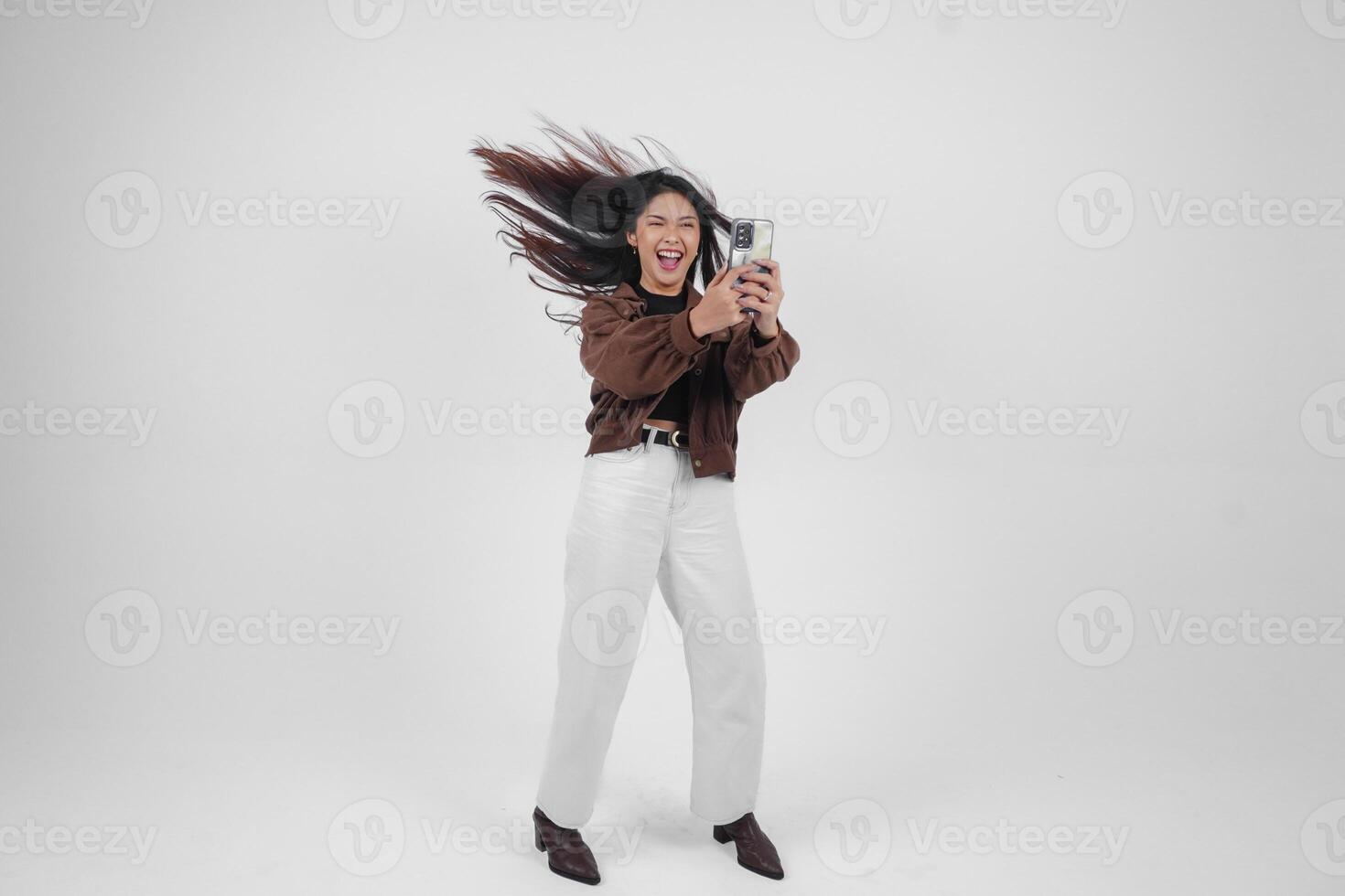 Cheerful Asian woman jumping and smiling in air while holding and looking at phone, feeling excited with a wide laugh on isolated white background photo