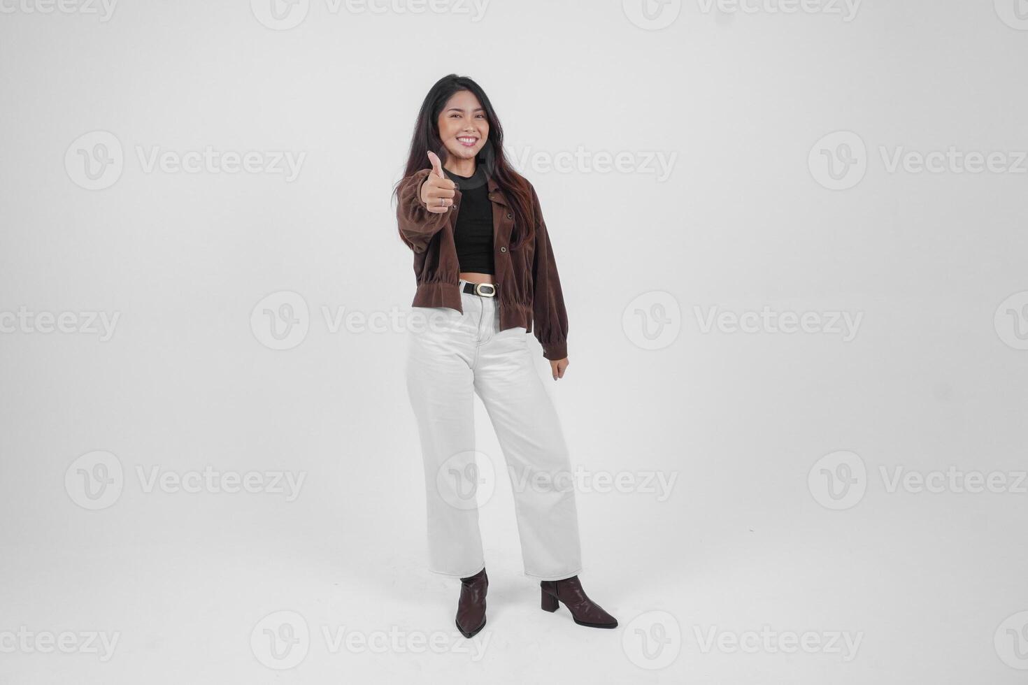 Attractive young Asian woman in brown jacket is gesturing thumbs up for approval, isolated by white background photo