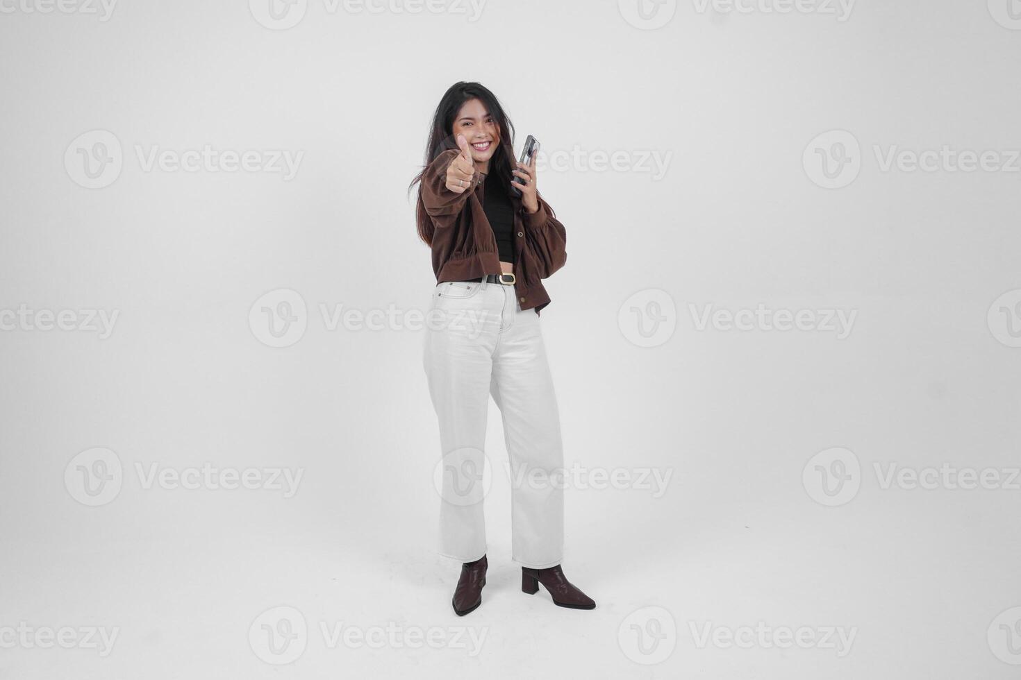 Cheerful young Asian woman showing thumbs up and holding phone using left hand, wearing casual clothes on isolated white background photo
