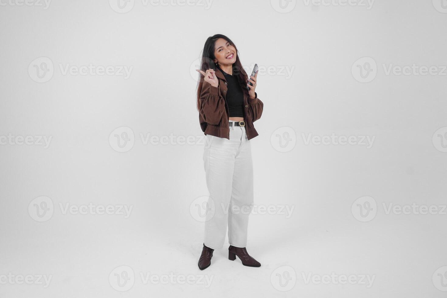 Smiling young Asian girl wearing brown jacket is standing while holding her phone and pointing to a copy space beside her. photo