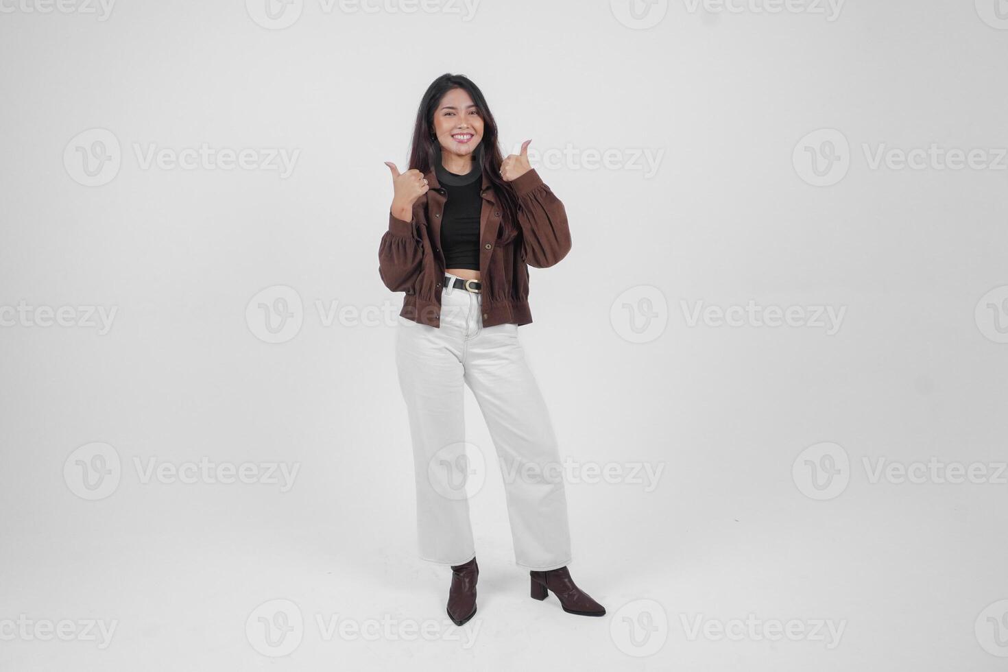 atractivo joven asiático mujer en marrón chaqueta es gesticulando pulgares arriba para aprobación, aislado por blanco antecedentes foto