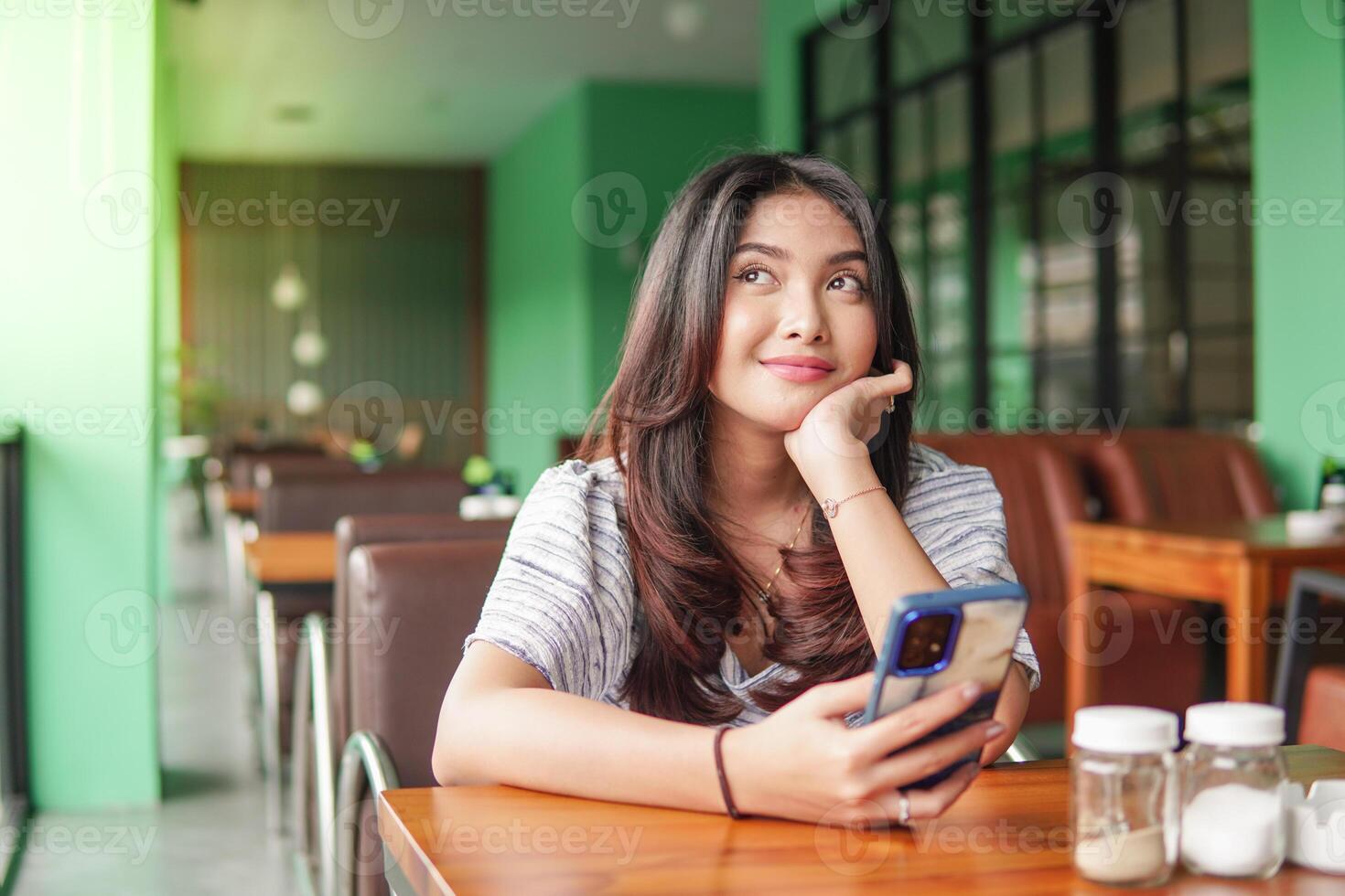 Thoughtful young Asian woman wearing dress sitting at a restaurant for a breakfast, holding a smartphone while propping her chin photo