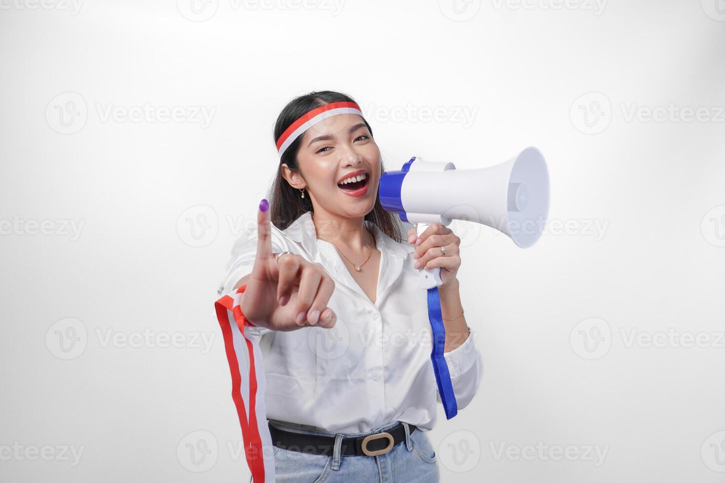 Energetic Indonesian woman in casual formal outfit wearing country flag headband while holding and shouting at megaphone, proudly showing little finger dipped in purple ink after voting for election photo