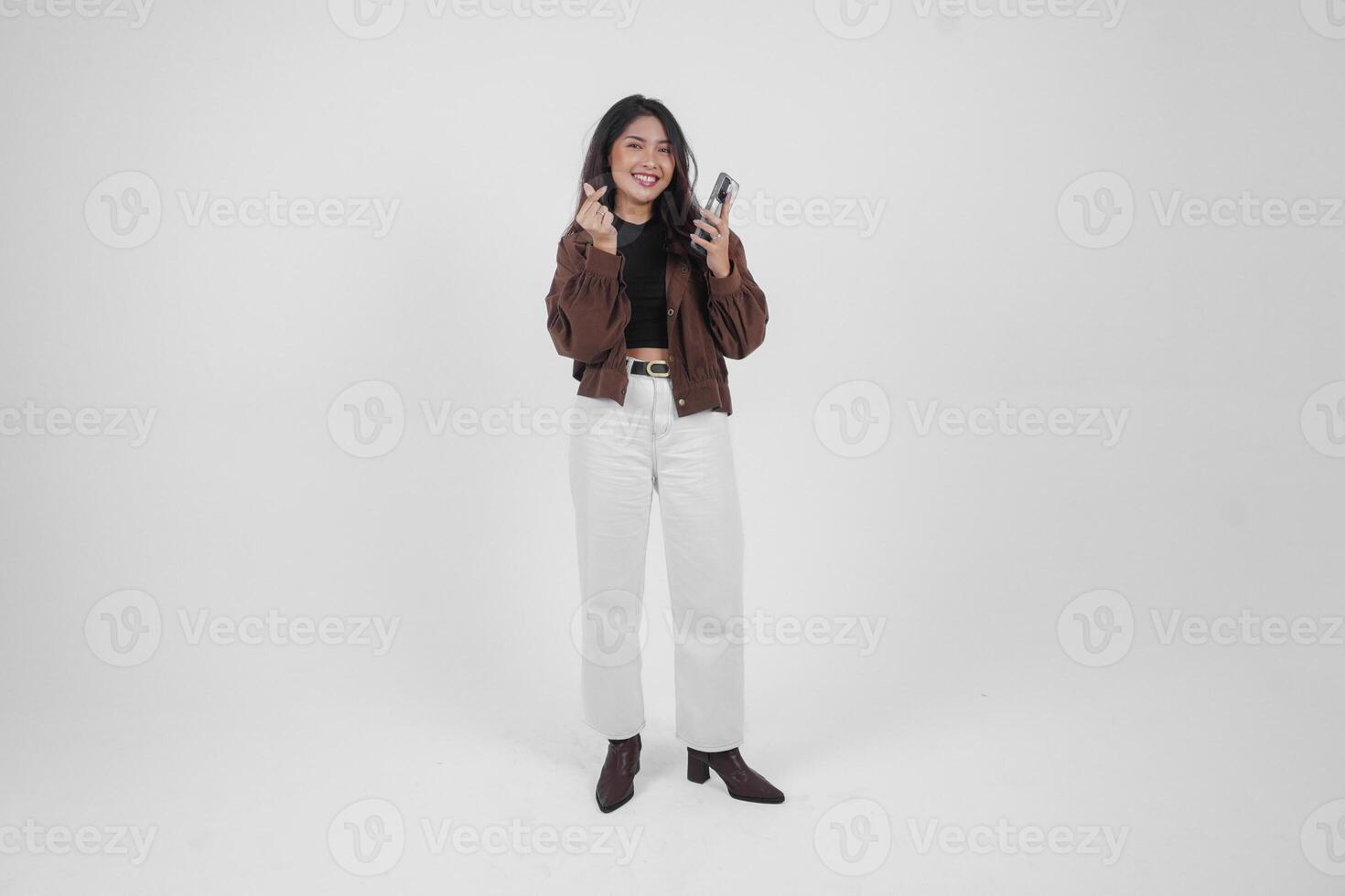 retrato lleno cuerpo de un joven asiático mujer haciendo amor corazón forma utilizando Derecha mano mientras participación teléfono utilizando izquierda mano en aislado blanco antecedentes foto