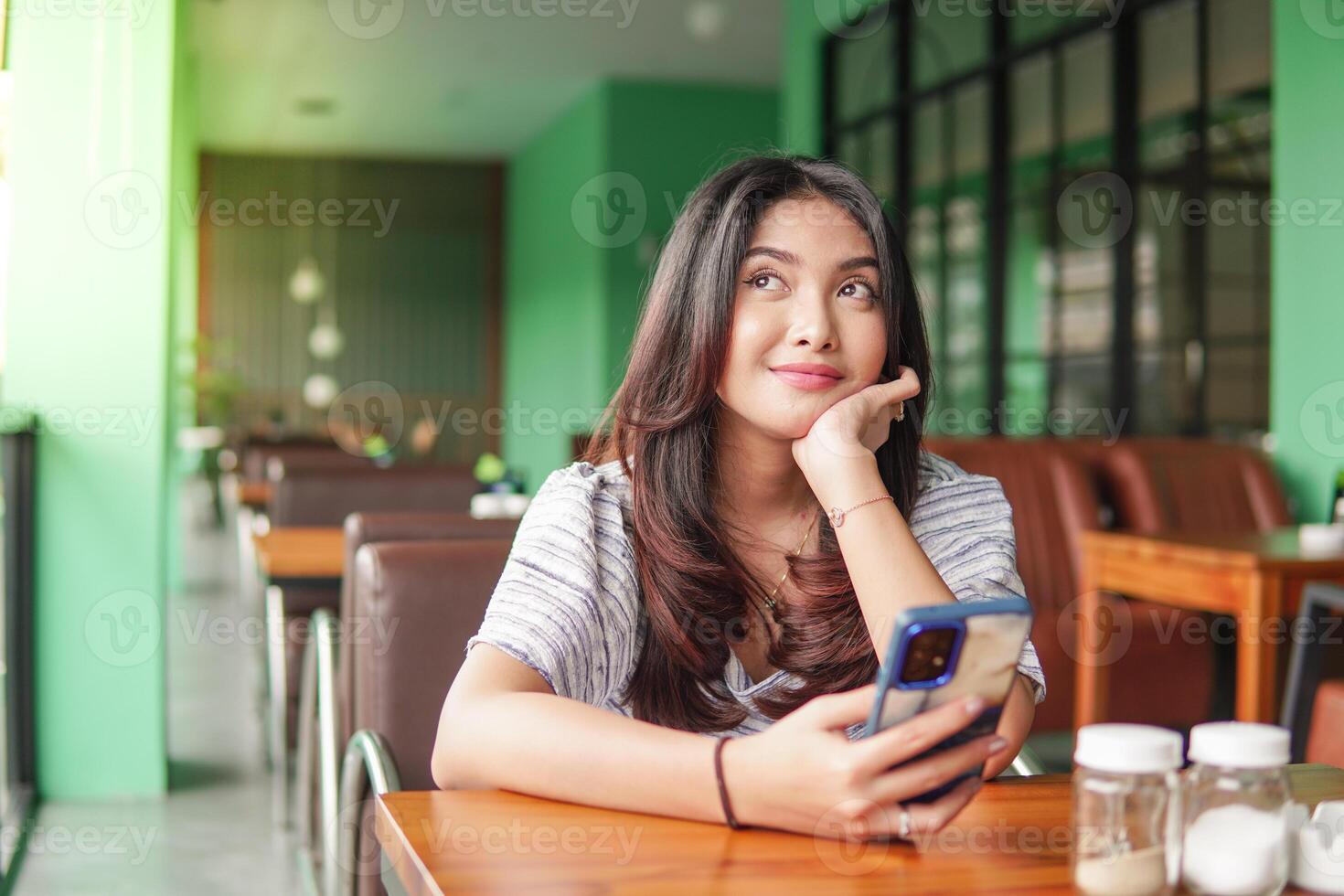 Thoughtful young Asian woman wearing dress sitting at a restaurant for a breakfast, holding a smartphone while propping her chin photo