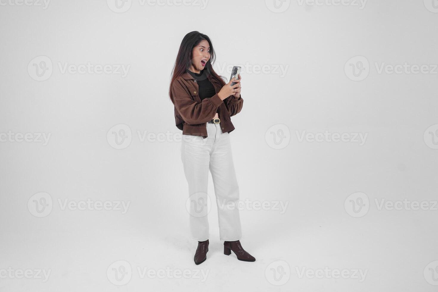 Portrait of a shocked Asian woman wearing casual outfit holding and showing her phone while her mouth wide open, standing on isolated white background photo