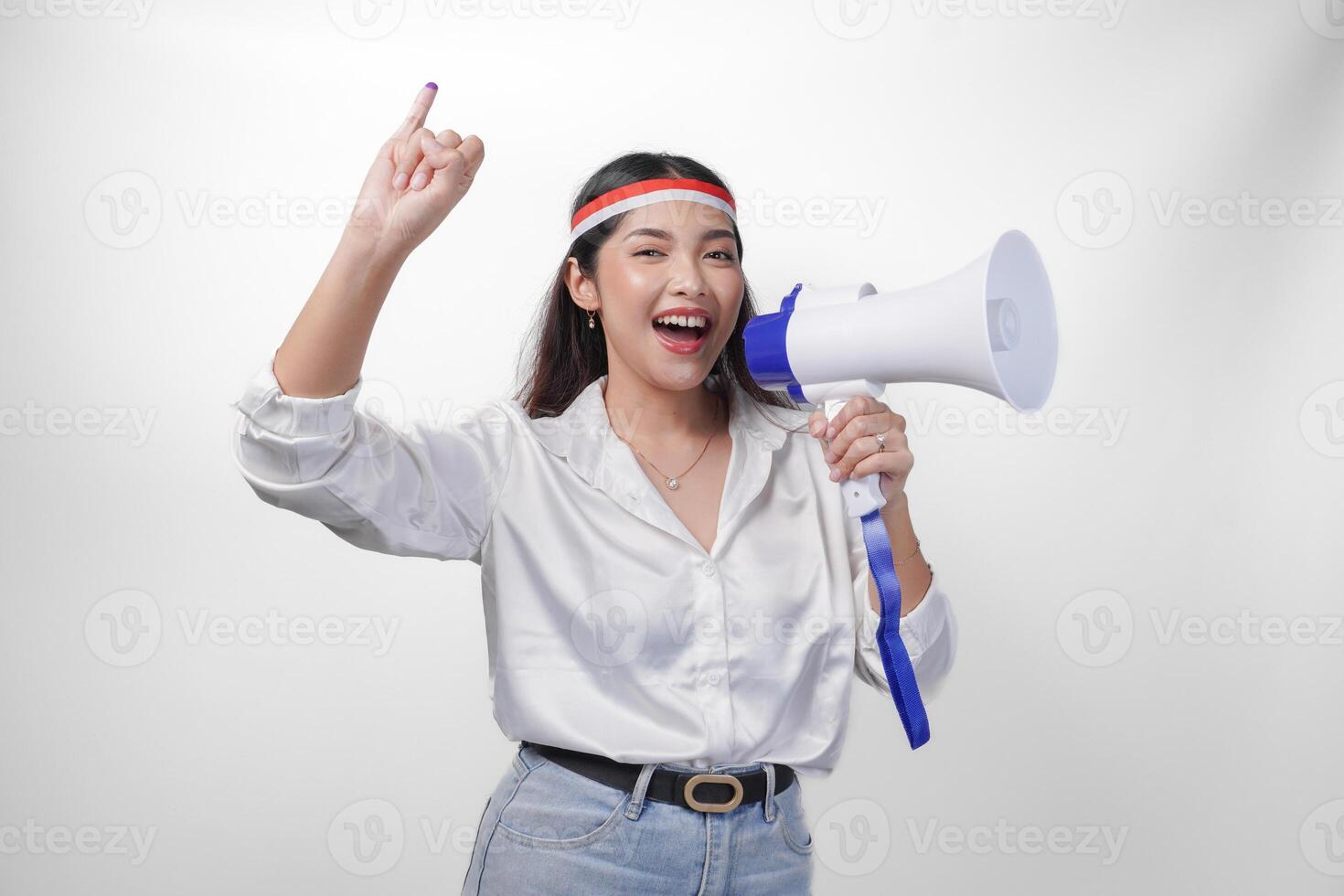 Energetic Indonesian woman in casual formal outfit wearing country flag headband while holding and shouting at megaphone, proudly showing little finger dipped in purple ink after voting for election photo
