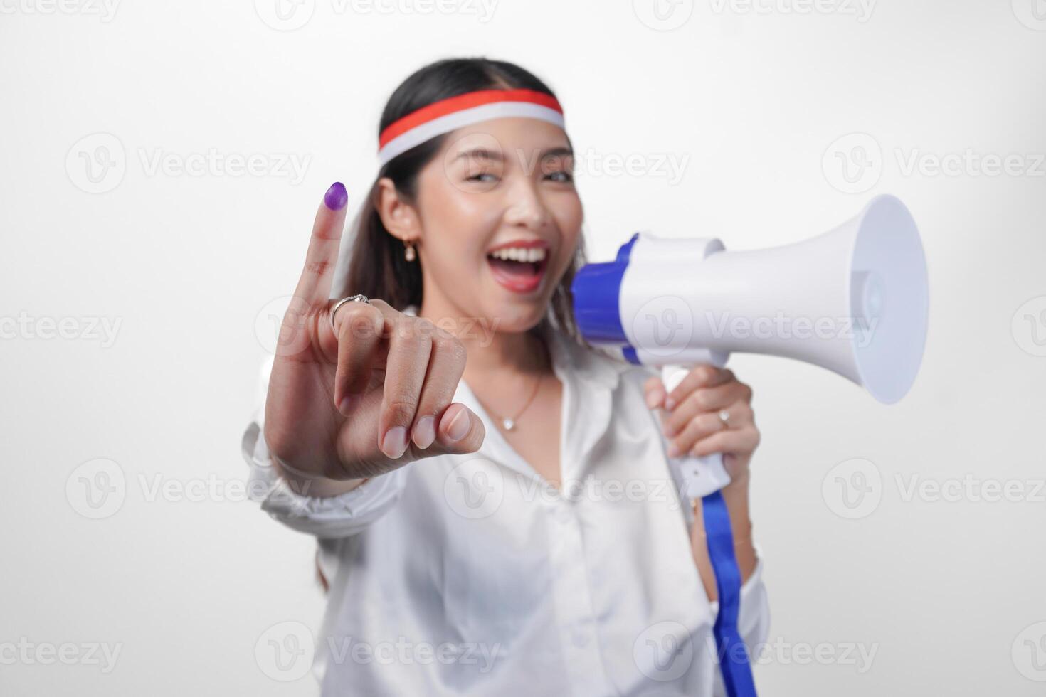 Energetic Indonesian woman in casual formal outfit wearing country flag headband while holding and shouting at megaphone, proudly showing little finger dipped in purple ink after voting for election photo