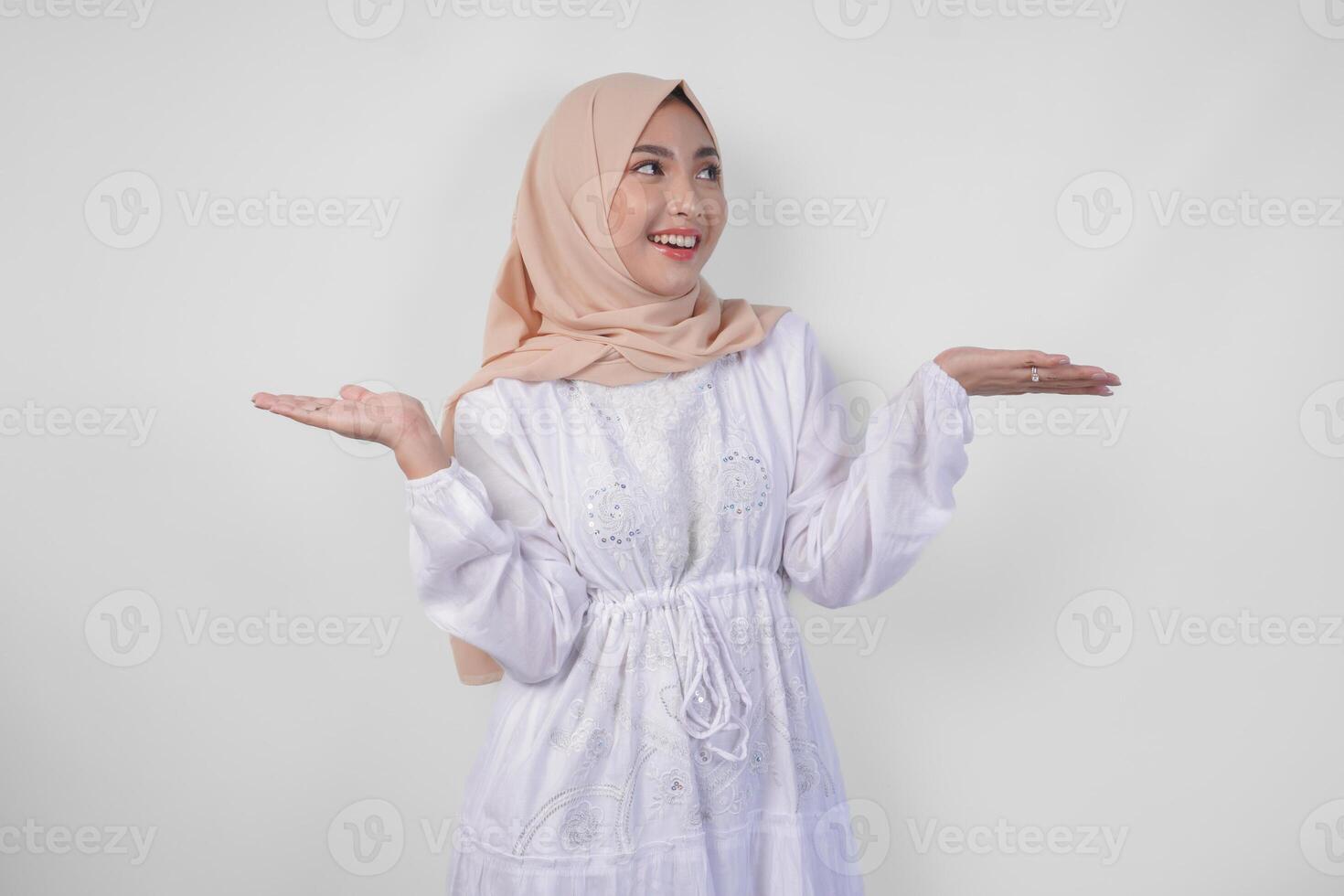 Happy Asian woman wearing hijab and white dress pointing up and presenting the option on copy space above, standing over isolated white background. Ramadan and Eid Mubarak advertisement concept photo