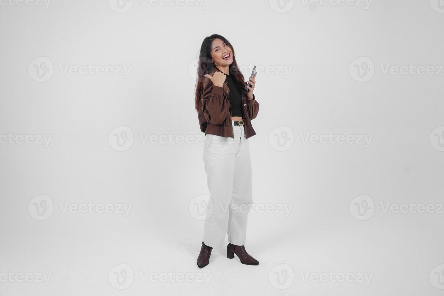 Smiling young Asian girl wearing brown jacket is standing while holding her phone and pointing to a copy space beside her. photo