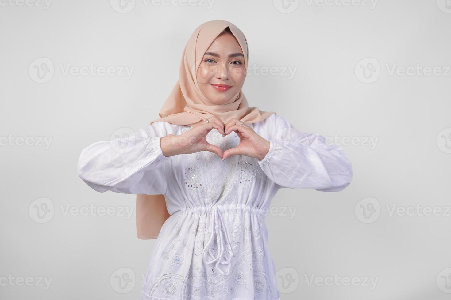 Young Asian Muslim woman in white dress and hijab making a heart love shape gesture with hands in various pose, isolated on white background photo