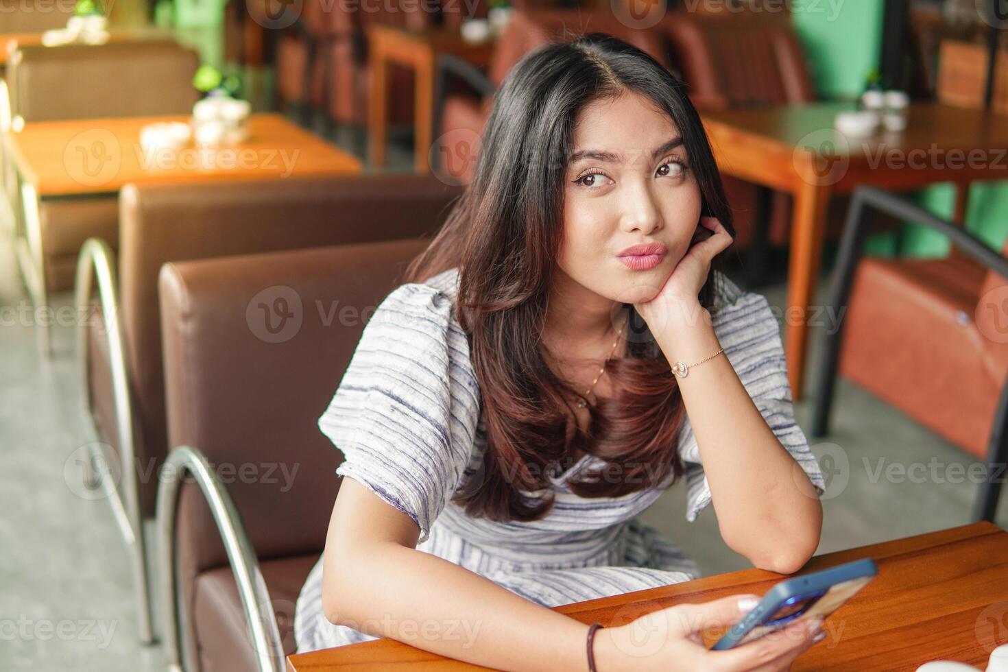 Thoughtful young Asian woman wearing dress sitting at a restaurant for a breakfast, holding a smartphone while propping her chin photo