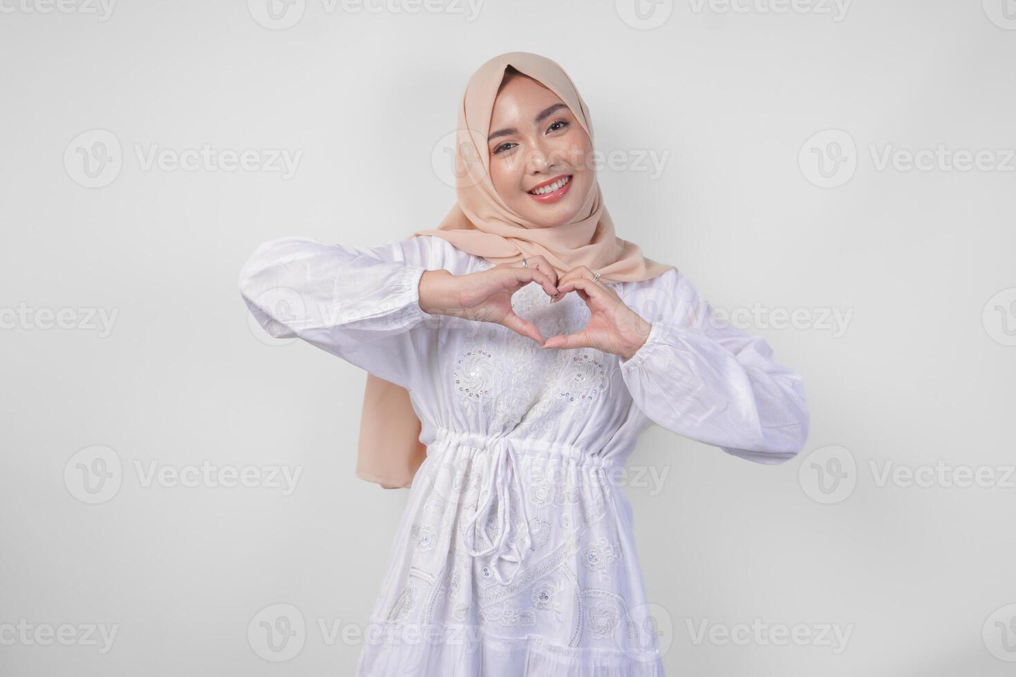 Young Asian Muslim woman in white dress and hijab making a heart love shape gesture with hands in various pose, isolated on white background photo
