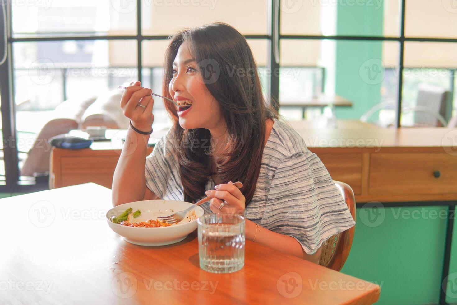 retrato de un emocionado joven asiático mujer vistiendo vestir sentado a un restaurante, comiendo y disfrutando desayuno con un alegre sonrisa foto