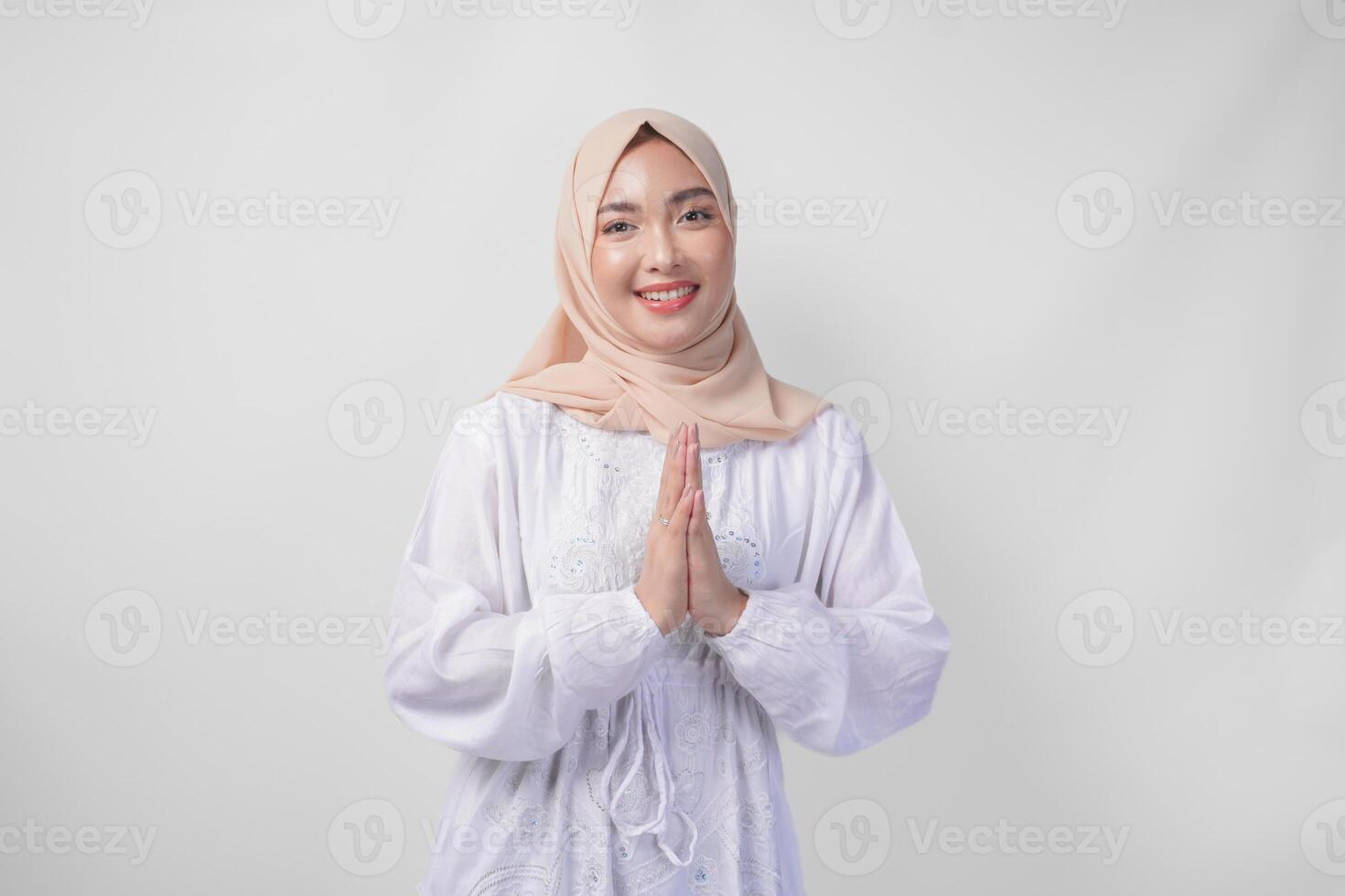 Beautiful Asian Muslim woman wearing white dress and hijab smiling while doing formal welcome or greeting gesture, standing over isolated white background photo