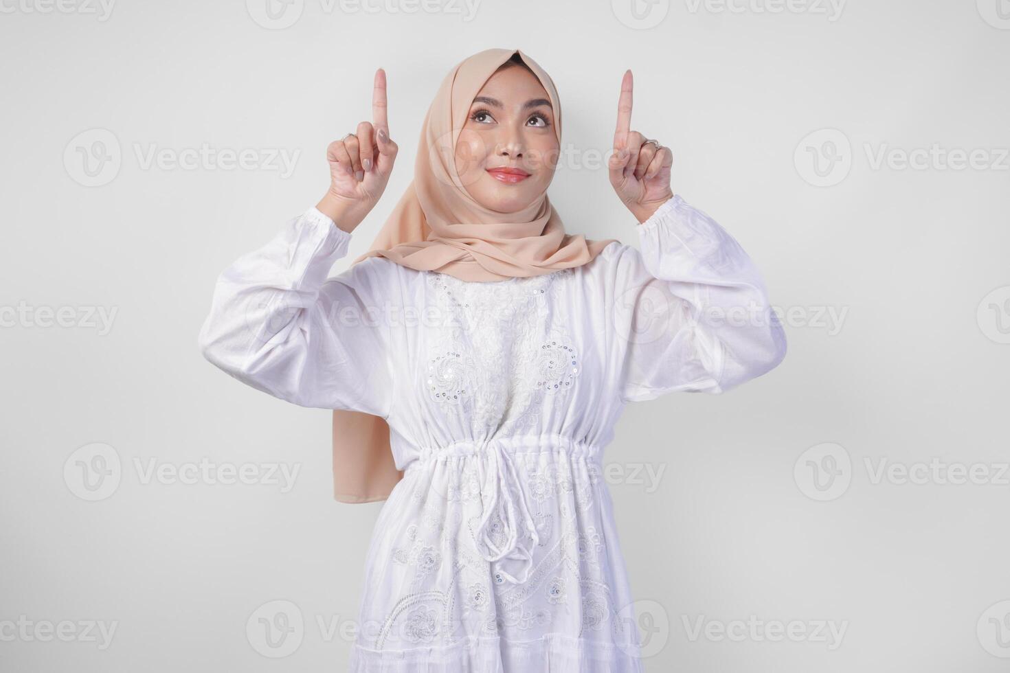 Happy Asian woman wearing hijab and white dress pointing up to the copy space above, standing over isolated white background. Ramadan and Eid Mubarak advertisement concept photo