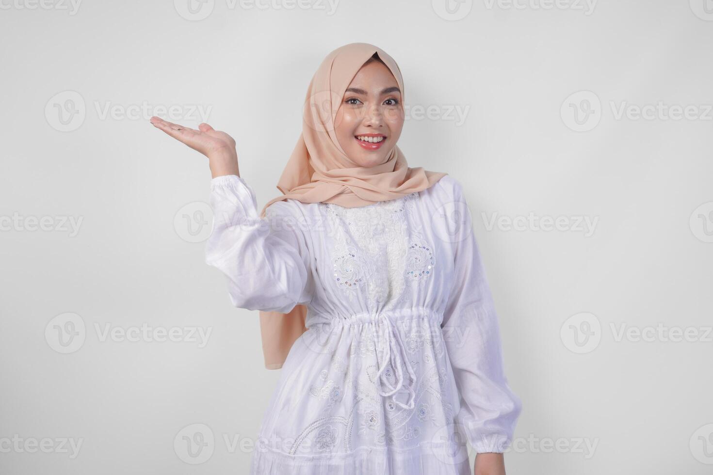 Smiling Asian woman wearing hijab and white dress pointing up to the copy space on her right side, standing over isolated white background. Ramadan and Eid Mubarak advertisement concept photo