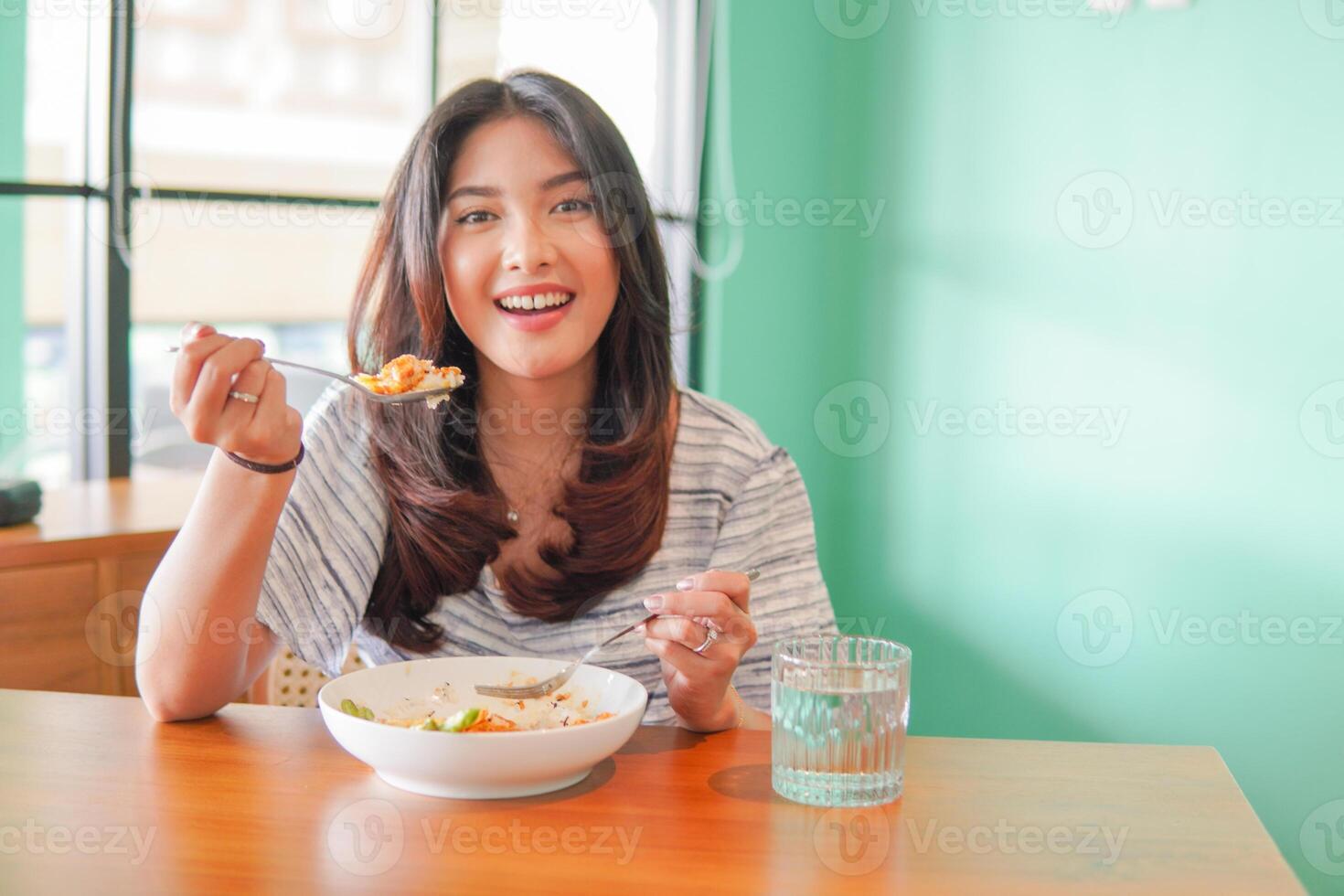 Portrait of an excited young Asian woman wearing dress sitting at a restaurant, eating and enjoying breakfast with a cheerful smile photo