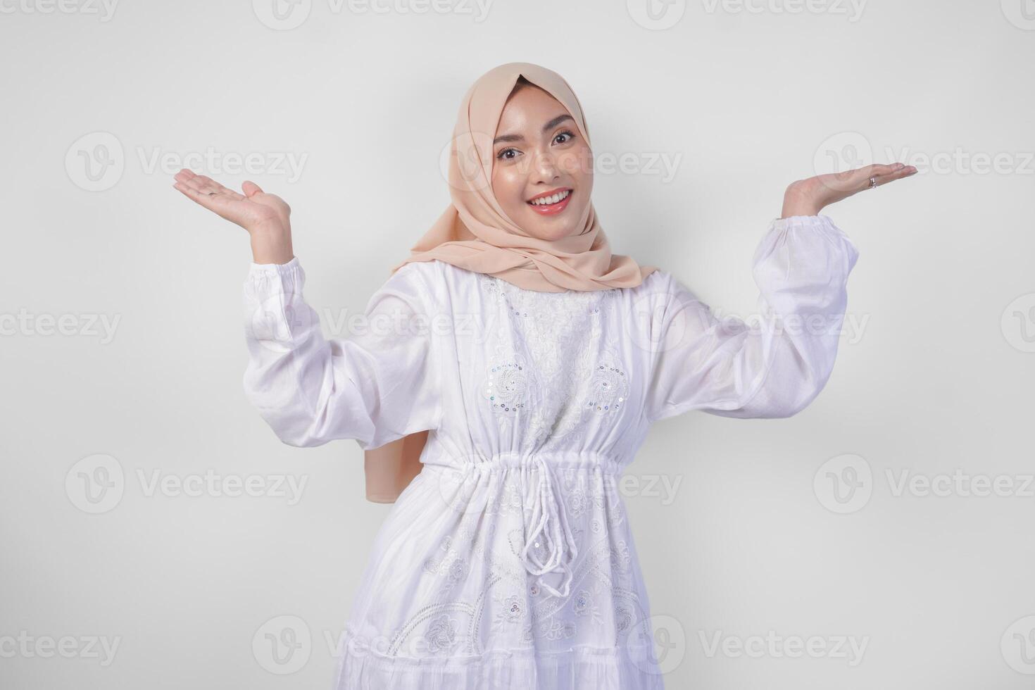 Happy Asian woman wearing hijab and white dress pointing up and presenting the option on copy space above, standing over isolated white background. Ramadan and Eid Mubarak advertisement concept photo