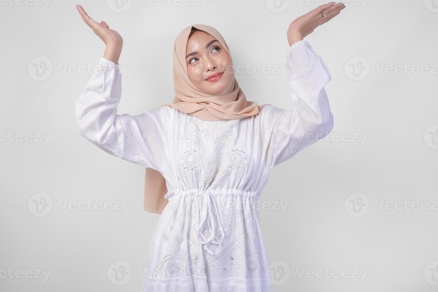 Happy Asian woman wearing hijab and white dress pointing up to the copy space above, standing over isolated white background. Ramadan and Eid Mubarak advertisement concept photo