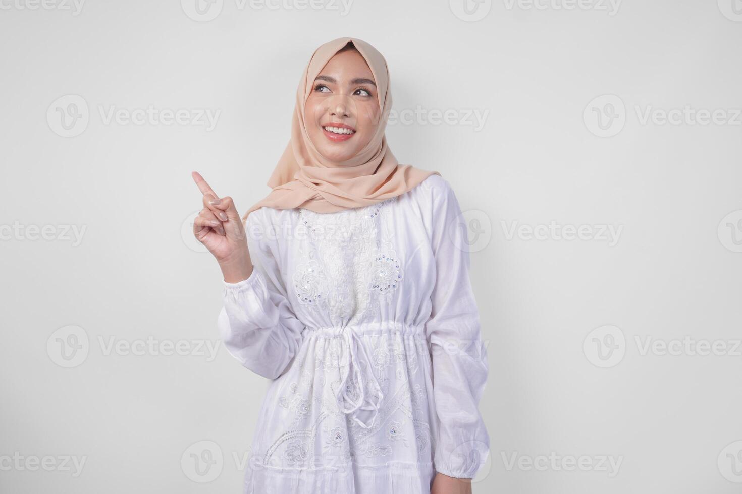 Smiling Asian woman wearing hijab and white dress pointing up to the copy space on her right side, standing over isolated white background. Ramadan and Eid Mubarak advertisement concept photo