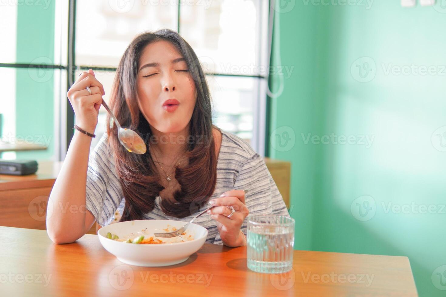 retrato de un emocionado joven asiático mujer vistiendo vestir sentado a un restaurante, comiendo y disfrutando desayuno con un alegre sonrisa foto
