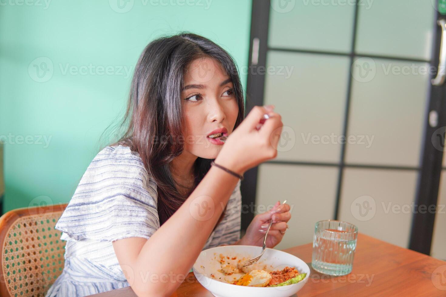 Portrait of an excited young Asian woman wearing dress sitting at a restaurant, eating and enjoying breakfast with a cheerful smile photo