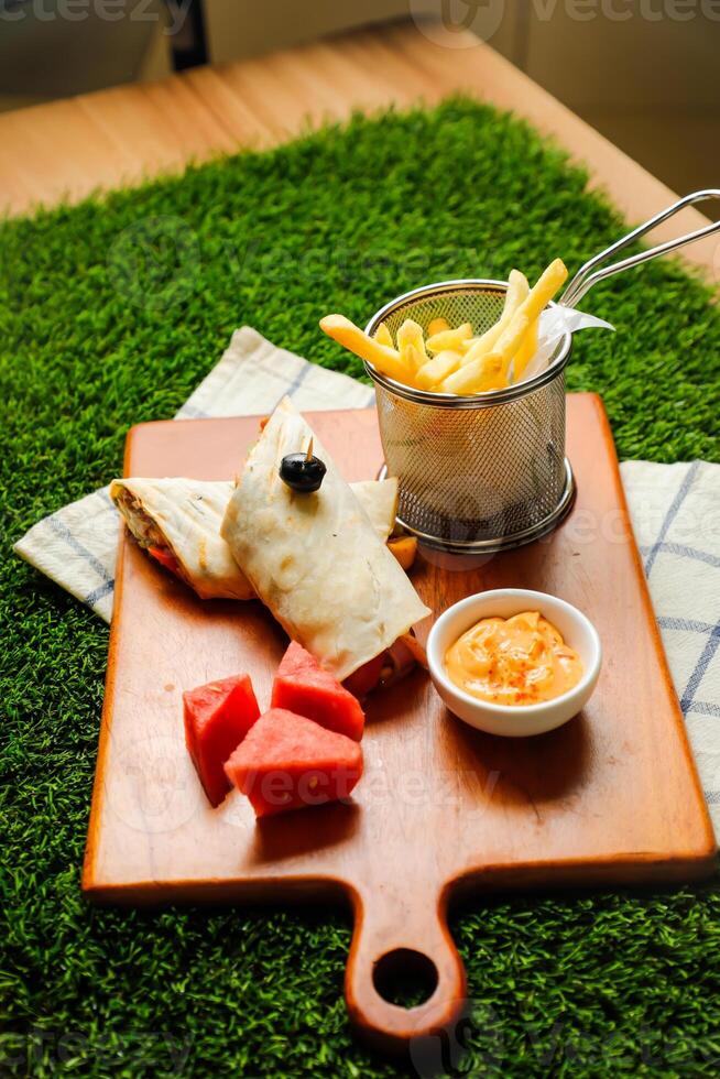 Chipotle Chicken Egg wrap roll filled with tomato, onion, cucumber, cheese with french fries, mayo dip and watermelon slice served on wooden board top view of fastfood appetizer photo